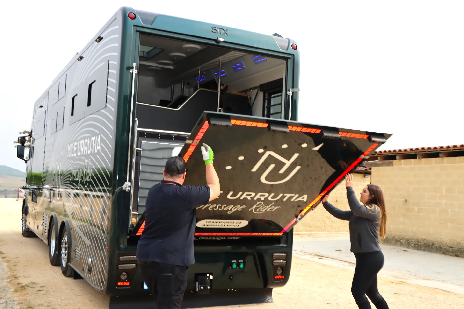 Dos personas abren la trampilla trasera de un gran camión de lujo para el transporte de caballos en un entorno exterior, demostrando la comodidad y eficacia del Borrador automático.