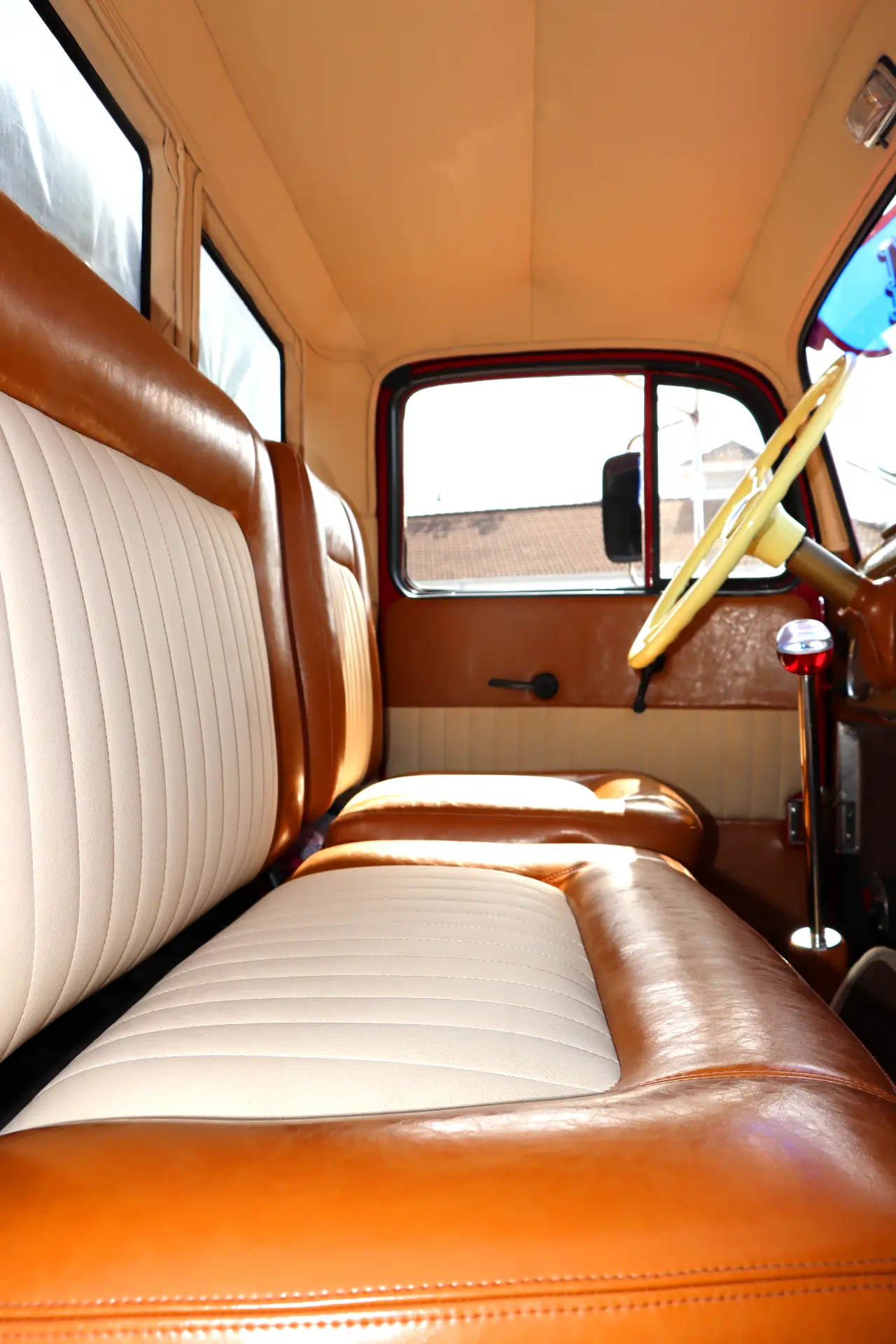 Interior de coche de época con asientos de cuero marrón y blanco, volante amarillo y luz solar brillante en el interior.