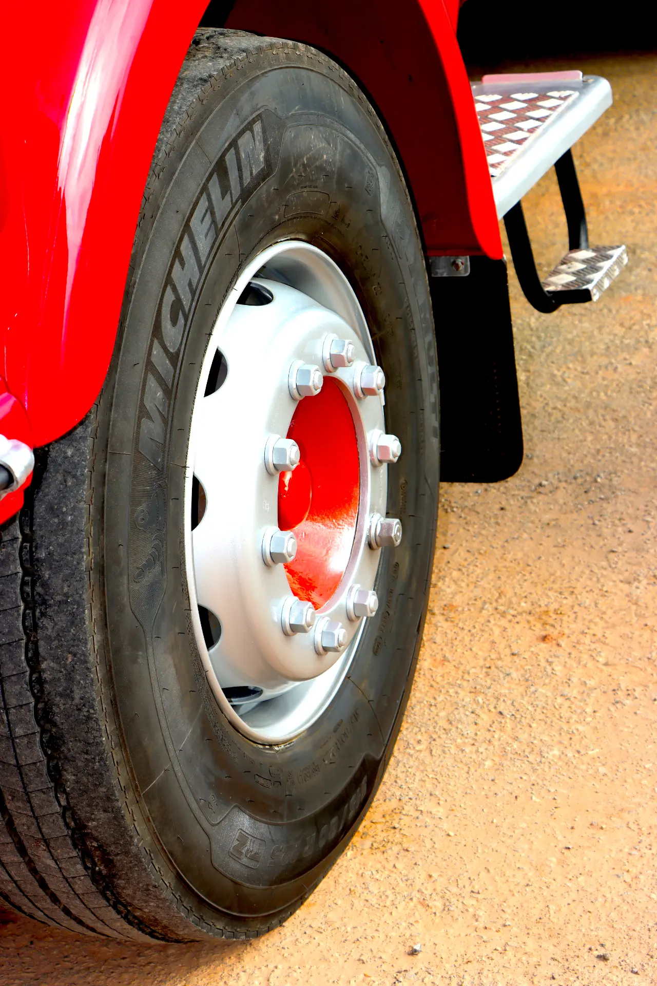 Primer plano de un gran neumático de camión Michelin en un vehículo rojo, aparcado en suelo arenoso.