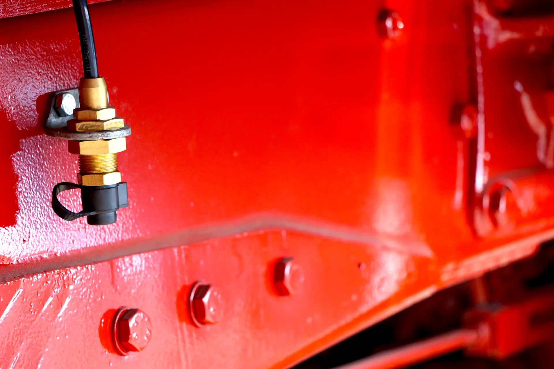 Primer plano de un sensor de latón fijado con pernos a una superficie metálica pintada de rojo.