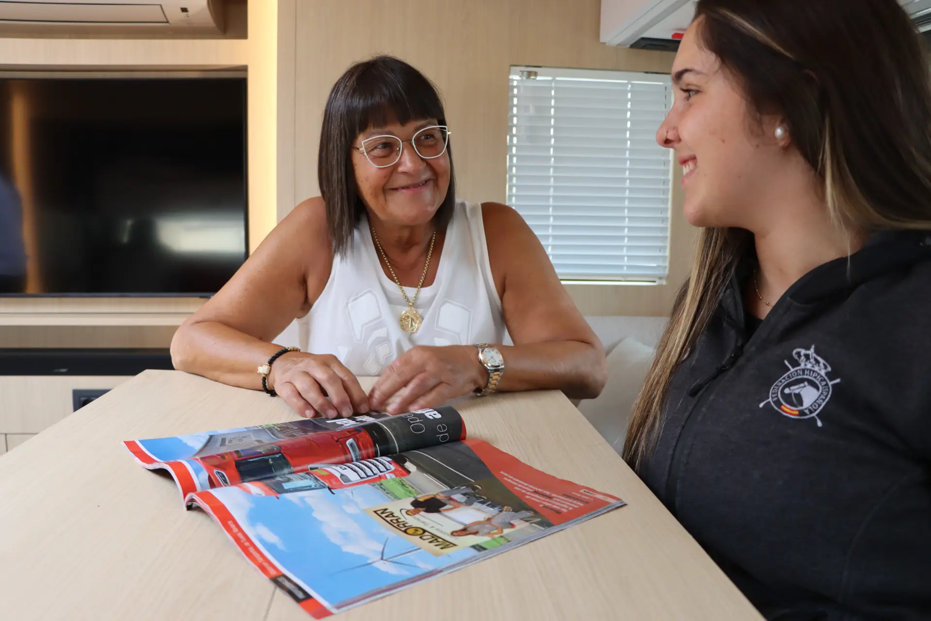 Dos mujeres sentadas a una mesa, sonriendo y hablando, con una revista abierta y un borrador automático delante.