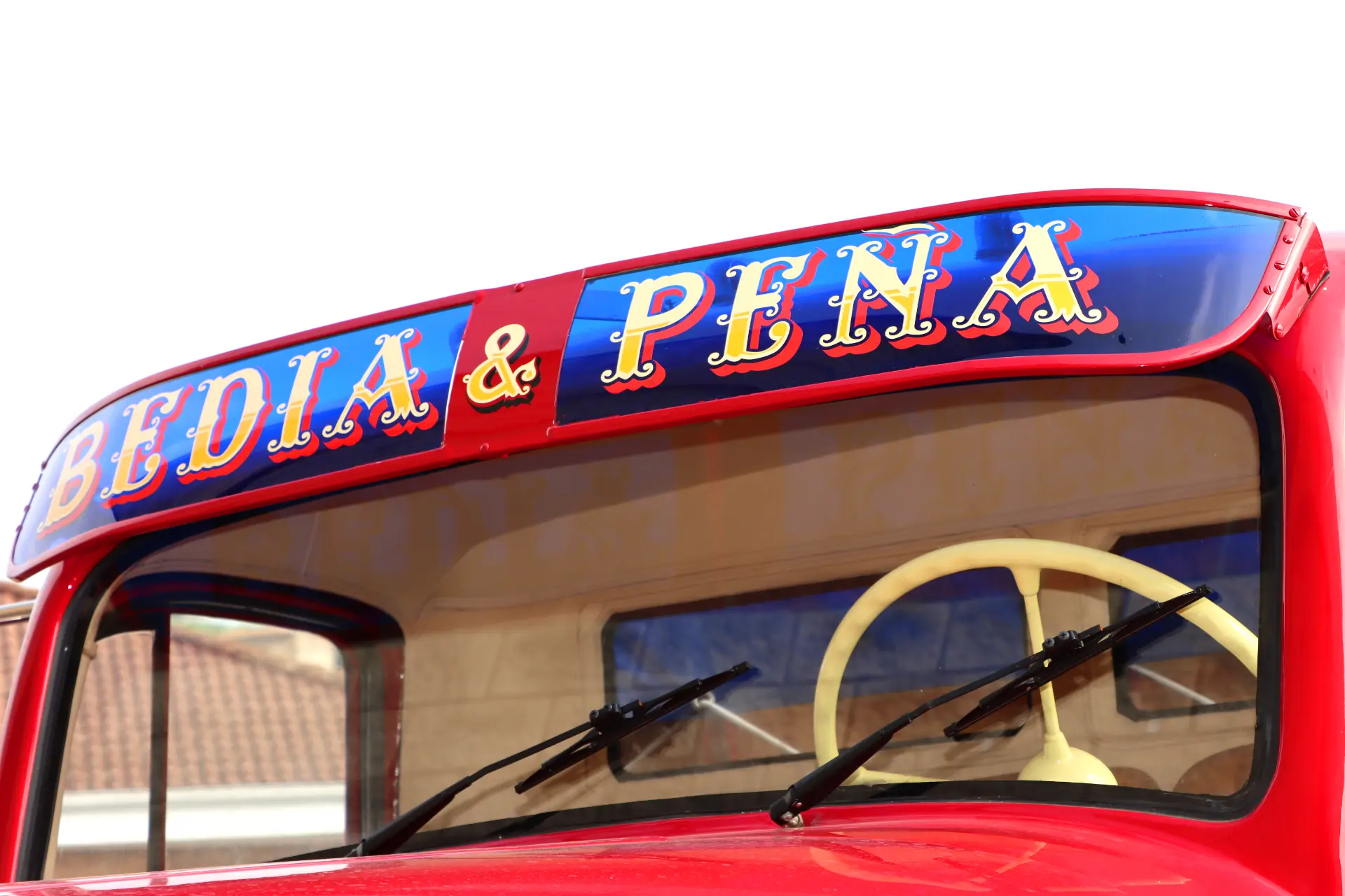 Vehículo rojo de época con "BEDIA & PENA" pintado en letras de colores sobre el parabrisas.