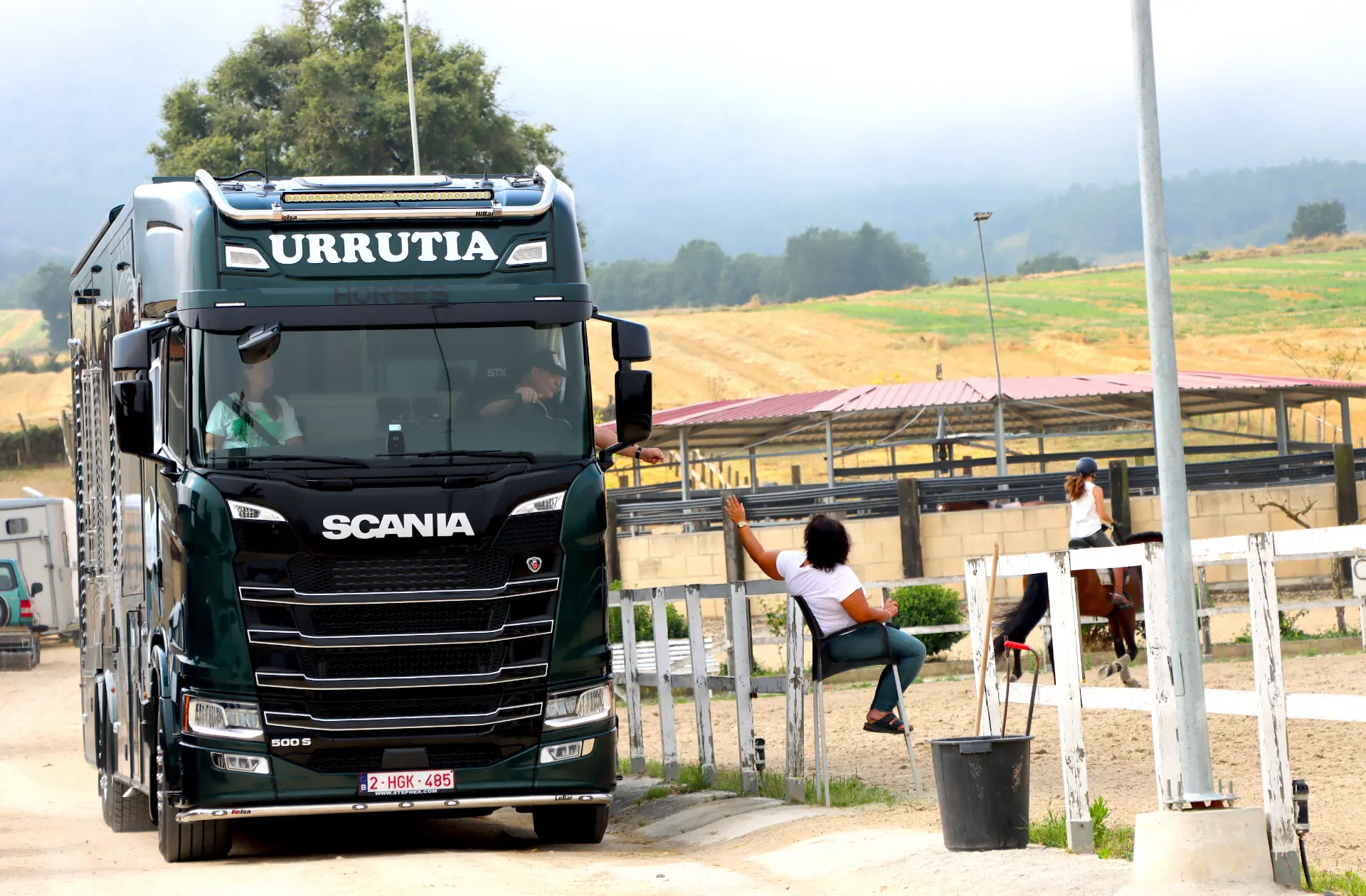 Un gran camión Scania, con un sistema Borrador automático, está aparcado cerca de unos establos de caballos, con personas y un caballo visibles al fondo.