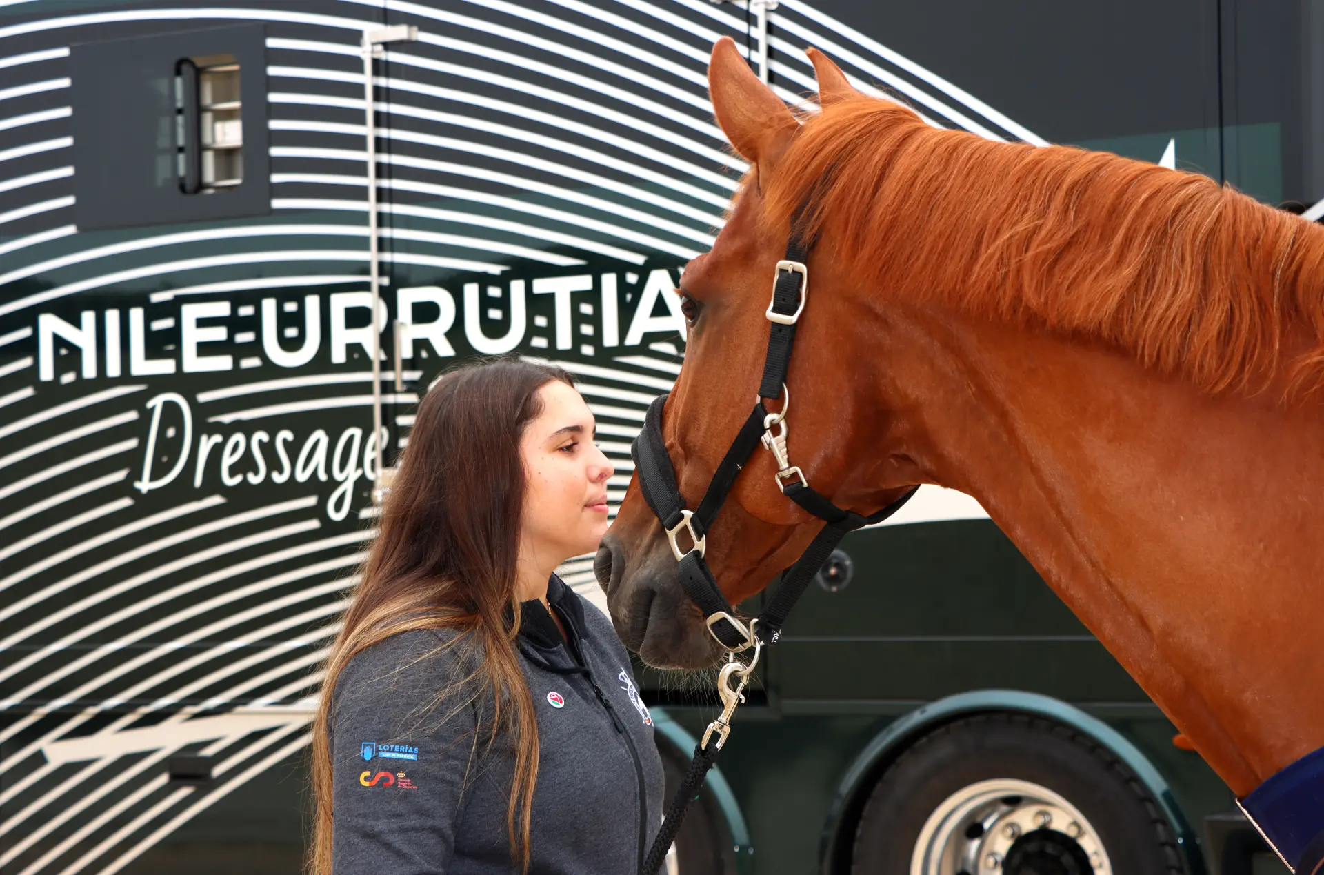 Mujer frente a un caballo marrón al aire libre, de pie junto a un vehículo con la palabra "Dressage" escrita en él.
