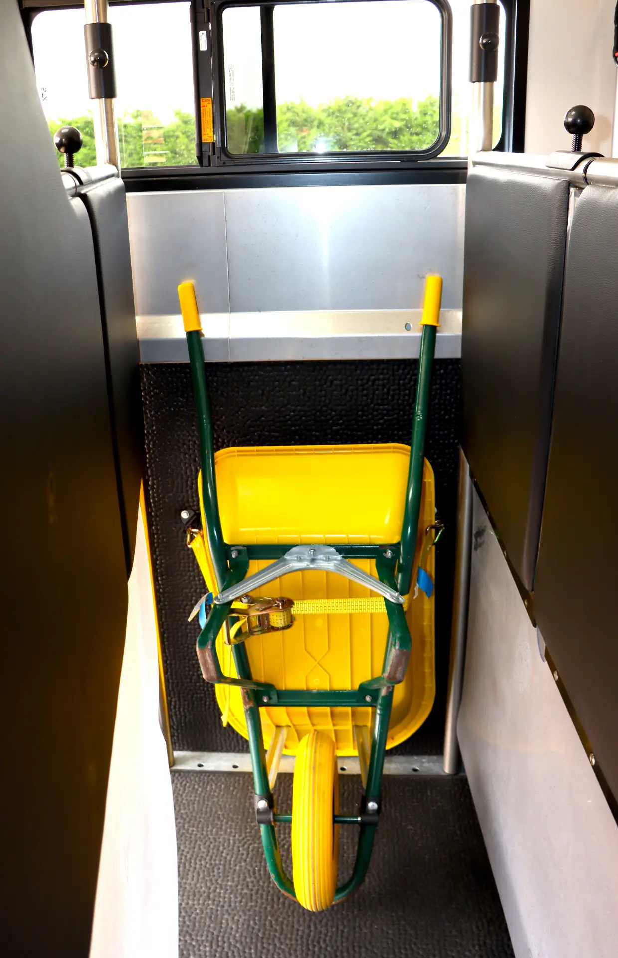 Una carretilla amarilla y verde, parecida en su forma a un borrador automático, está amarrada en posición vertical dentro del pasillo de un vehículo entre asientos negros.