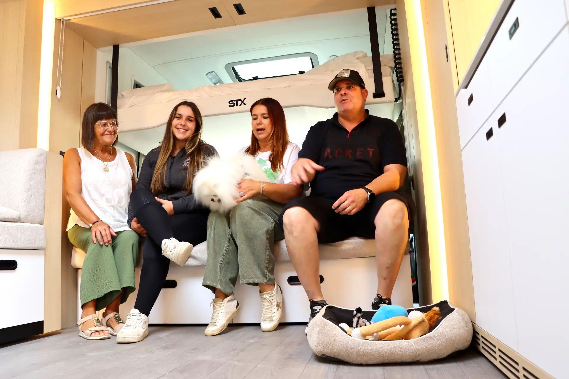 Cuatro personas y un esponjoso perro blanco se sientan en el interior de una furgoneta camper, con una cama para perros y juguetes en el suelo.