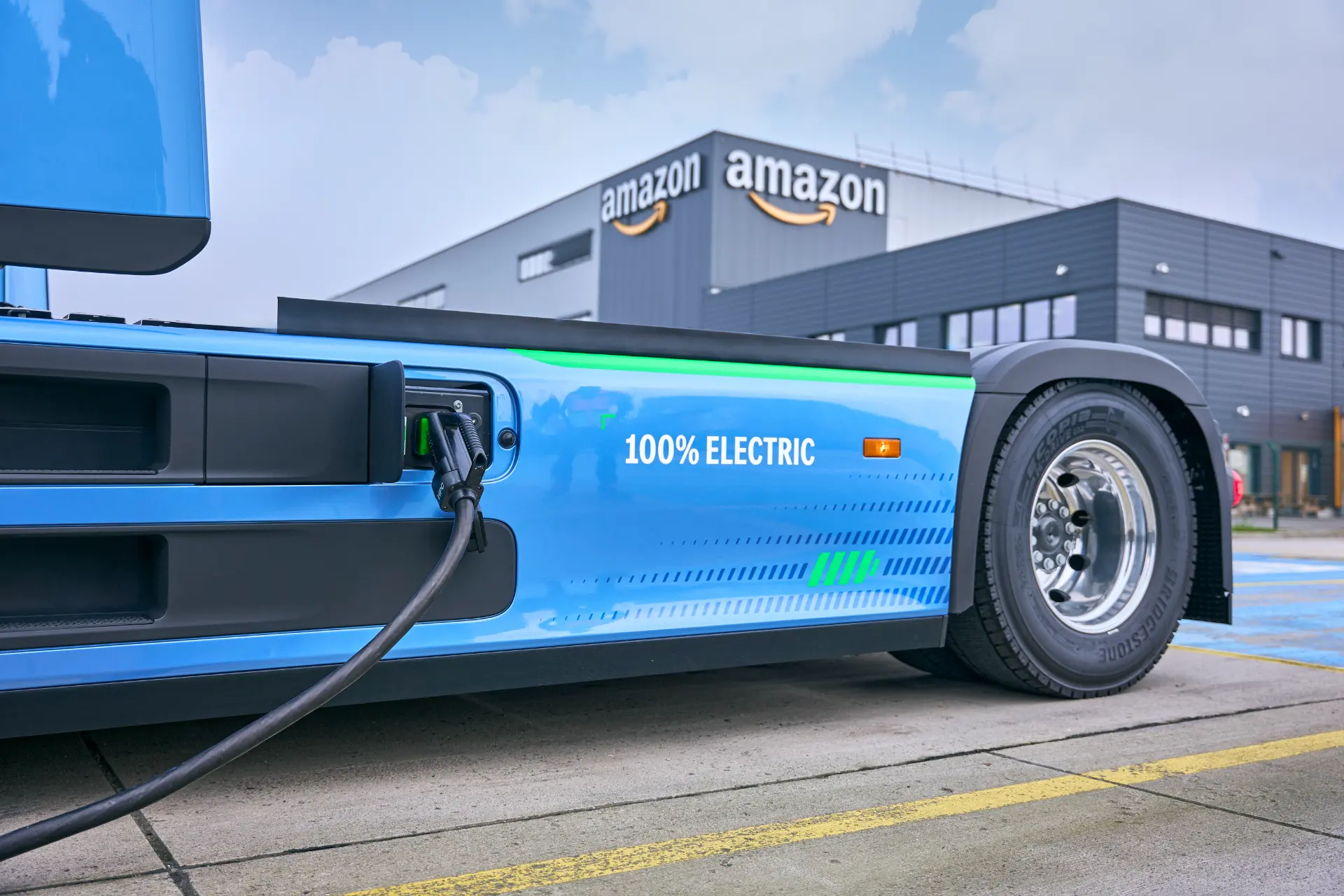 Un camión eléctrico azul cargando frente a un almacén de Amazon, con las leyendas "100% eléctrico" y "Borrador automático" escritas en su lateral.