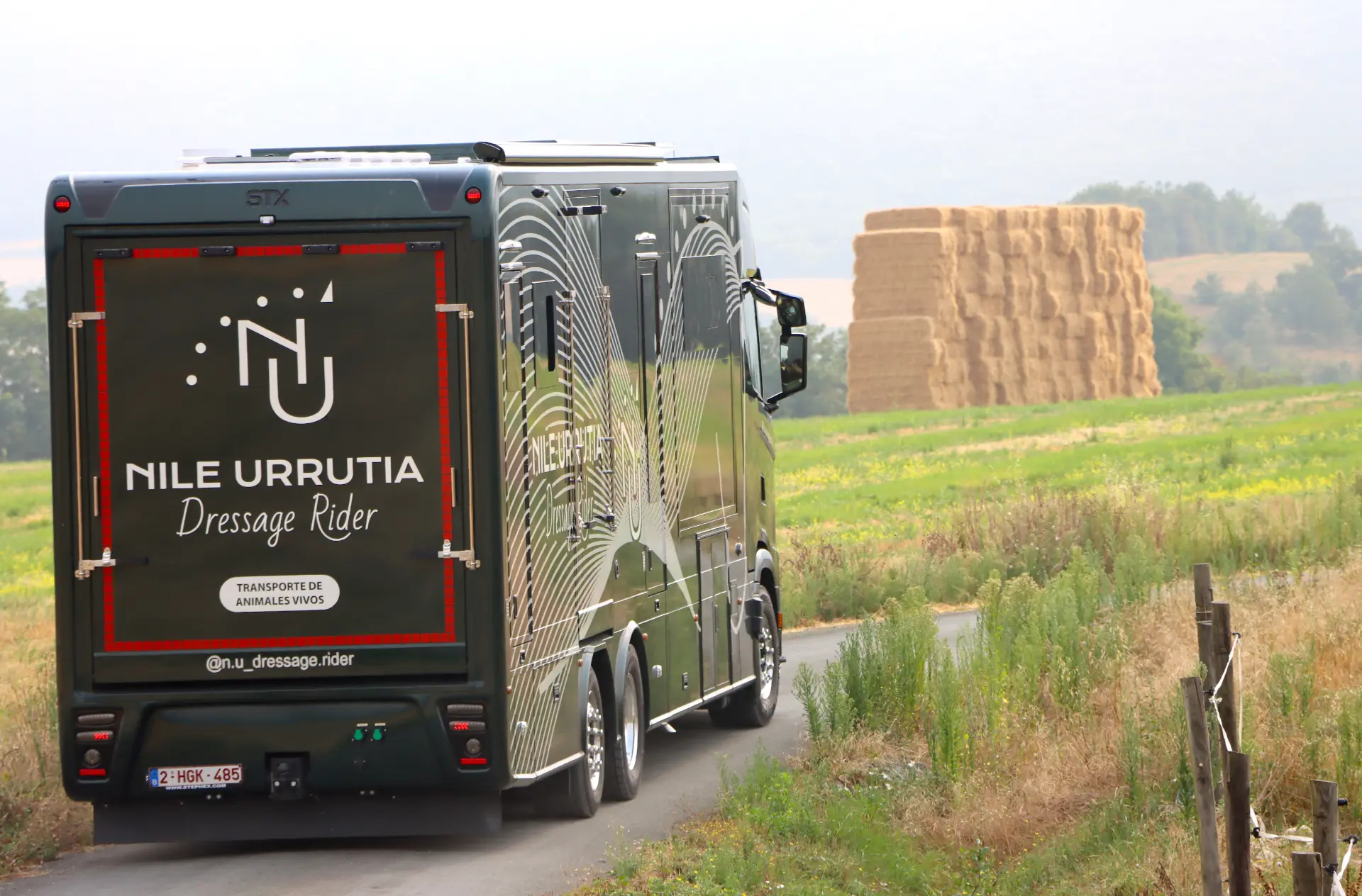 Un gran camión de transporte de caballos circula por una carretera rural junto a un campo con pacas de heno apiladas.