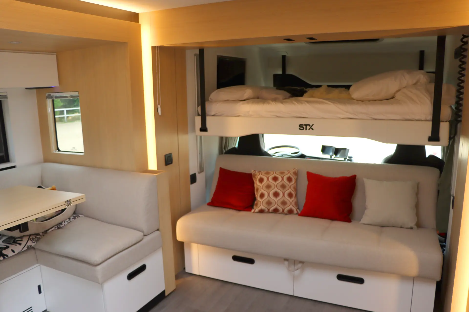Moderno interior de autocaravana con estación de borrador automático, con una litera sobre un sofá, almohadas decorativas y una zona de comedor a la izquierda.