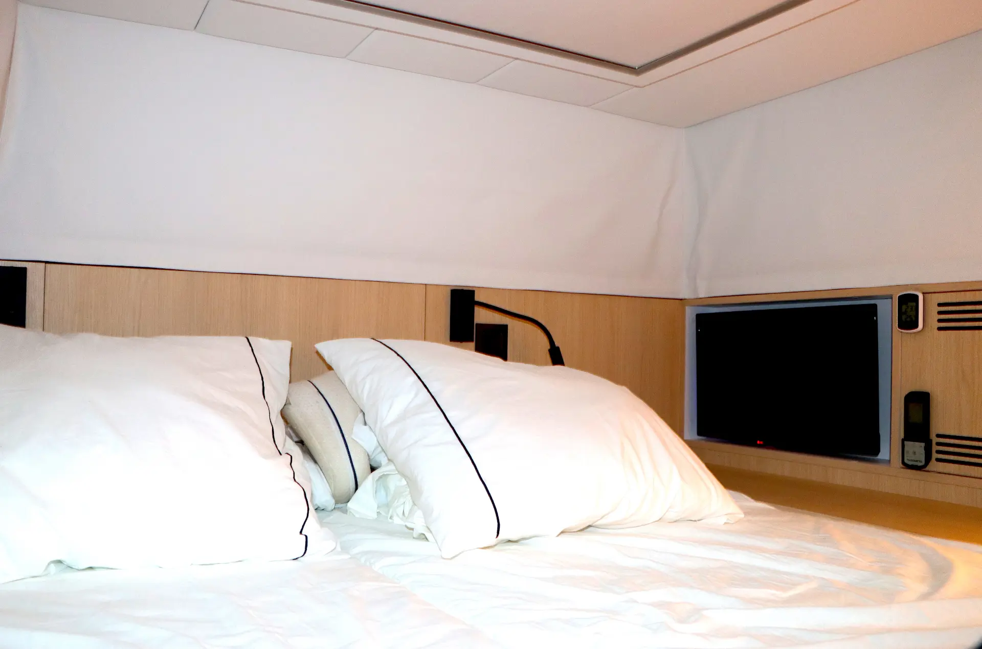 Una cama bien hecha con almohadas blancas, una luz de lectura y un televisor montado en la pared crean un ambiente moderno en la habitación, perfecto para relajarse o trabajar en su Borrador automático.