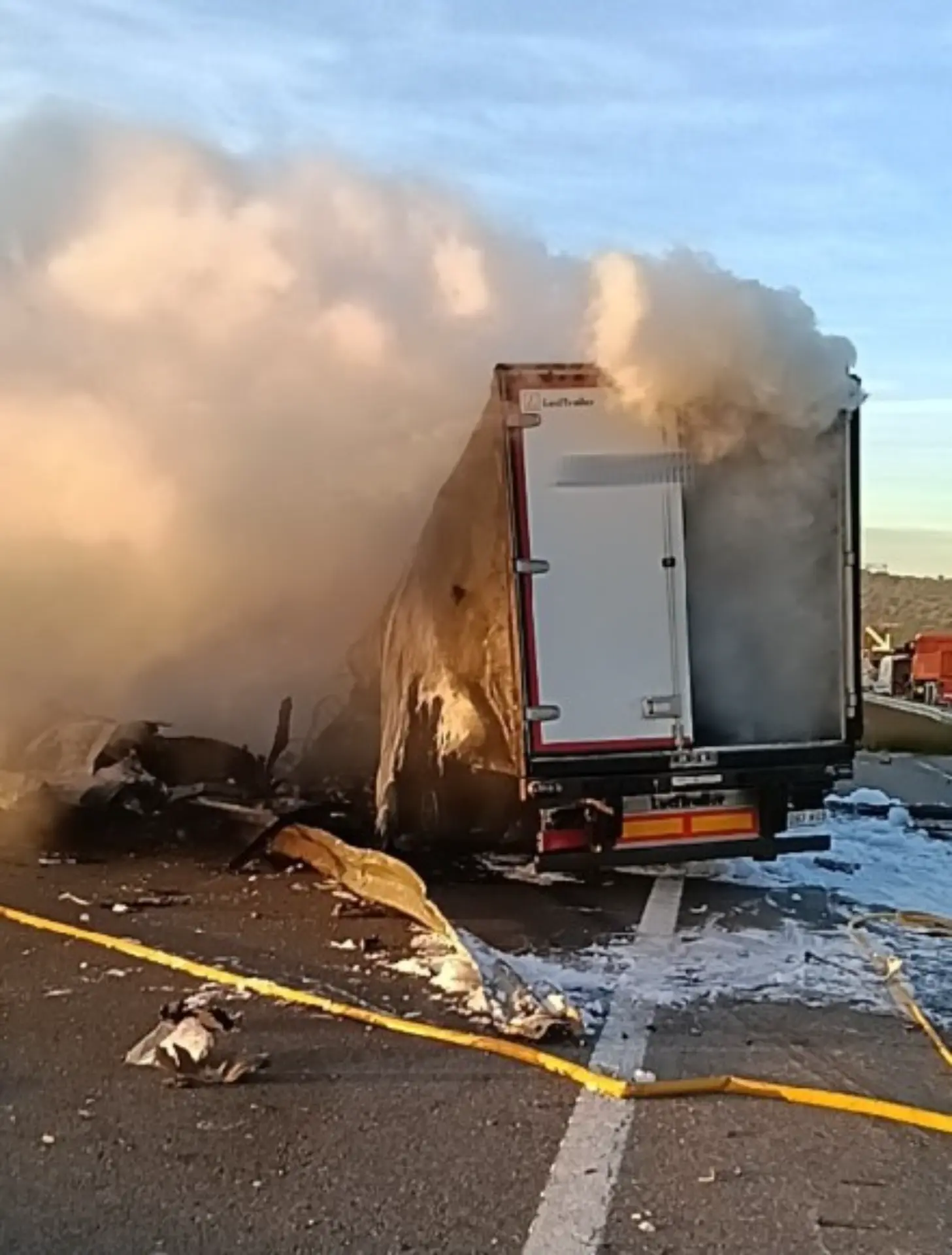 Un camión en llamas con humo espeso saliendo, parcialmente destruido, en la carretera AP-7 tras un accidente múltiple, con cinta de precaución cerca.