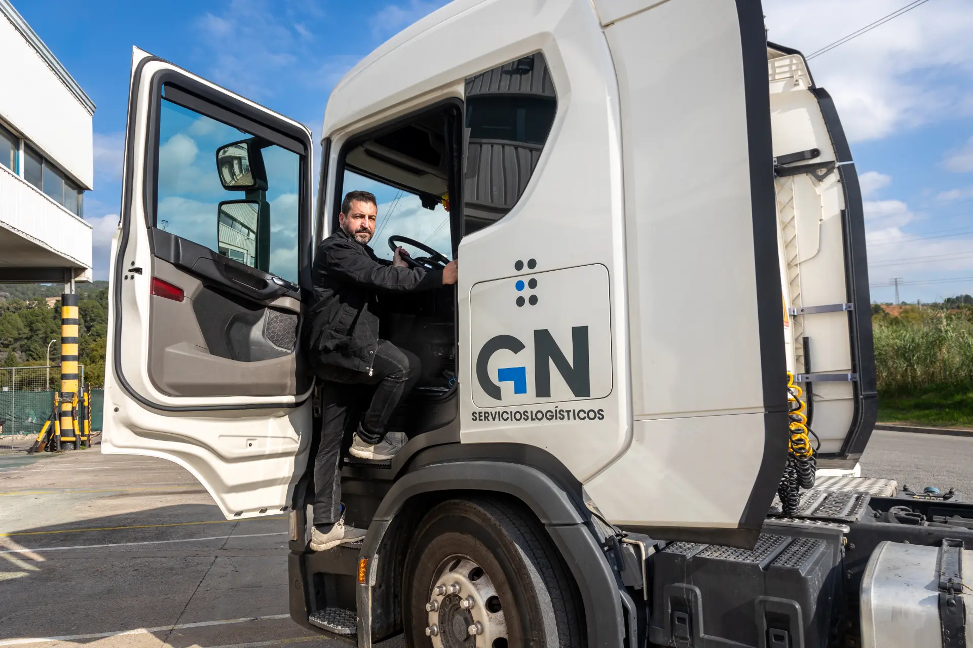 Hombre de pie en las escaleras de un camión, sujetando la puerta junto a un camión blanco de "GN Servicios Logísticos" al aire libre, comprobando su borrador automático antes de iniciar la ruta.
