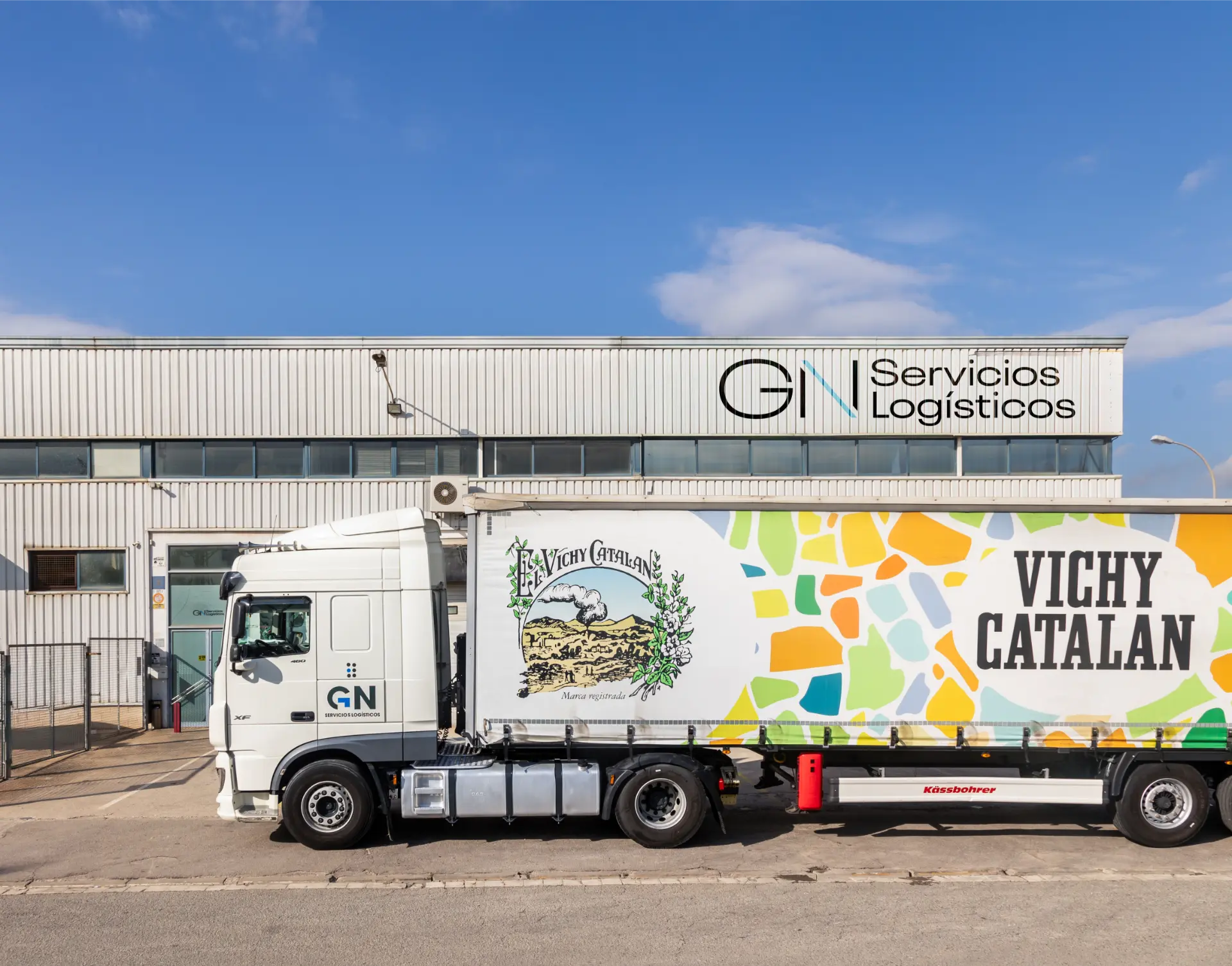 Un camión blanco de GN Servicios Logísticos con un colorido remolque vichy catalán, marcado como "Borrador automático", aparcado junto a un almacén.