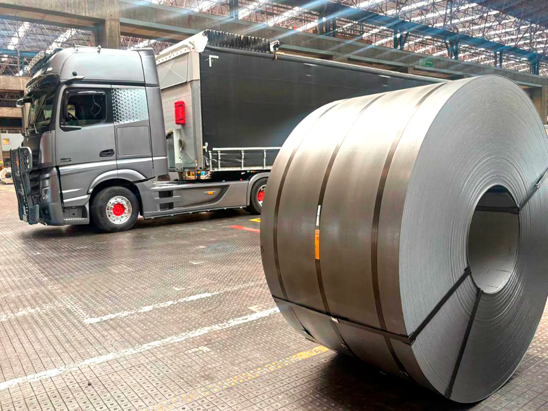 Una gran bobina de metal, que forma parte de un proceso automatizado con un sistema de borrador automático, se encuentra delante de un camión gris aparcado en el interior de una nave industrial.