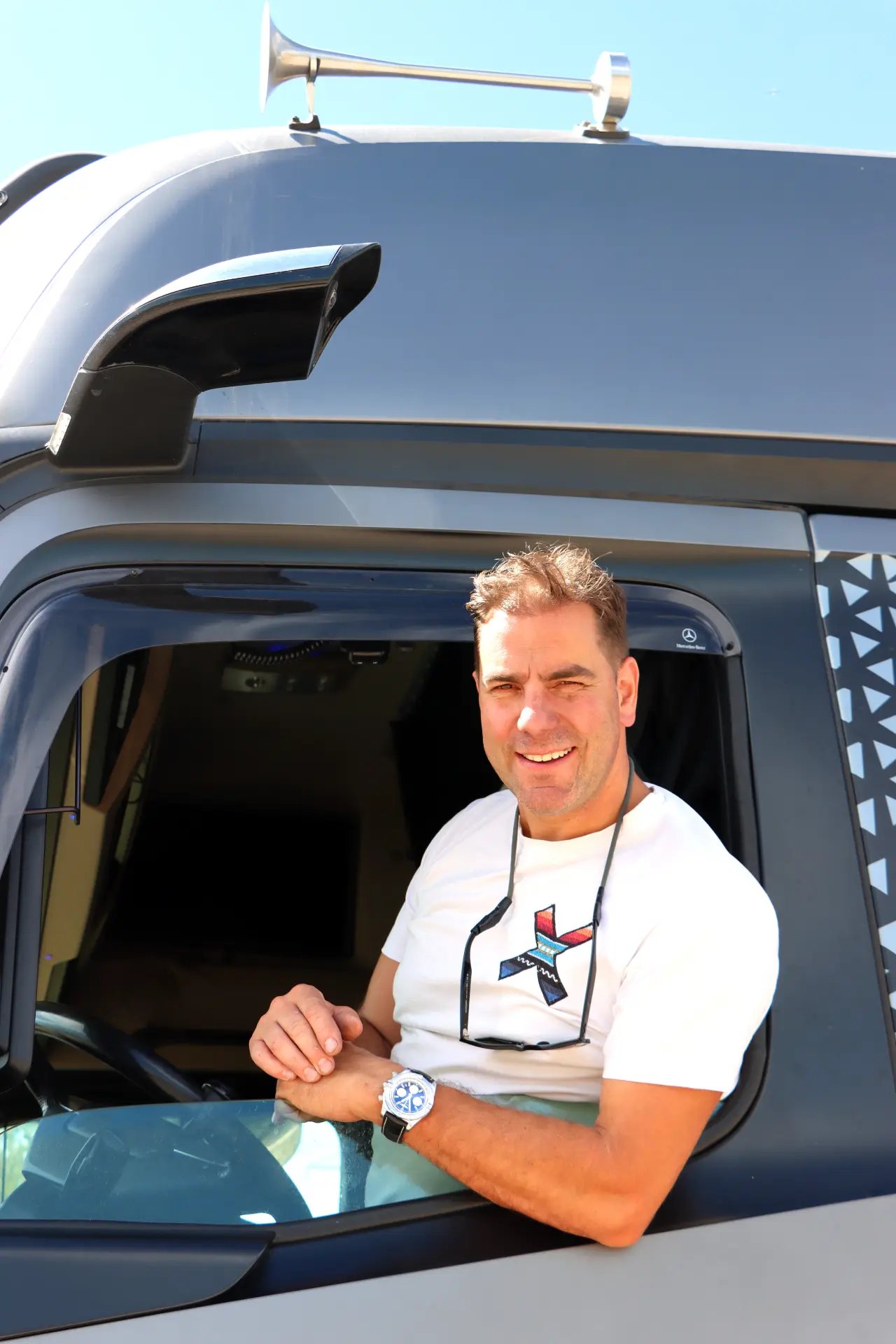 Un hombre sonriente con camiseta blanca se asoma a la ventanilla de un camión en un día soleado, sosteniendo un borrador automático.