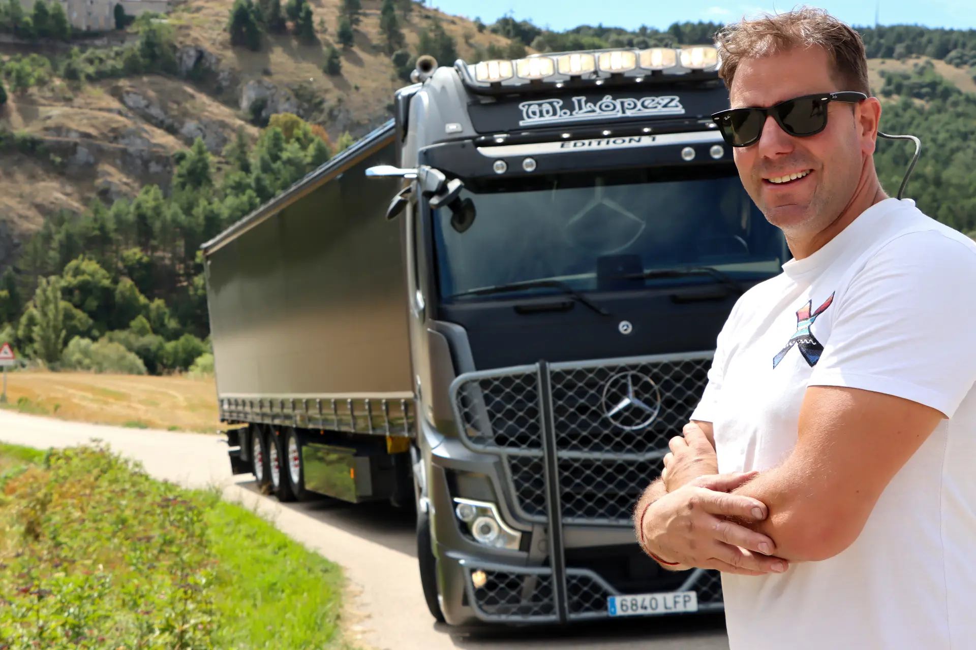 Hombre con gafas de sol de pie y sonriendo delante de un semirremolque Mercedes-Benz negro en una carretera rural, como si posara para un anuncio de borrador automático.