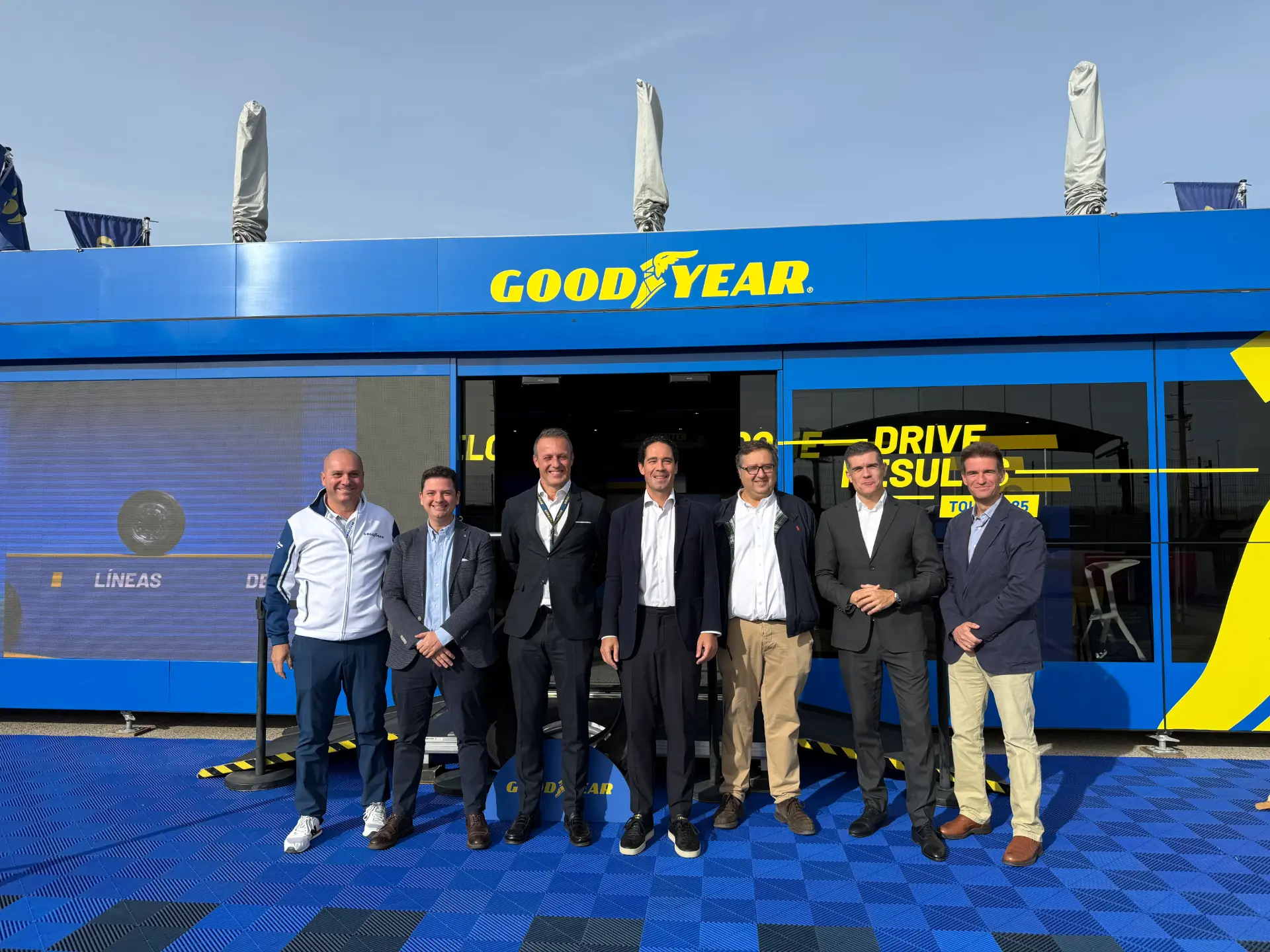 Siete hombres en traje de negocios frente a un edificio azul de Goodyear en un día soleado.