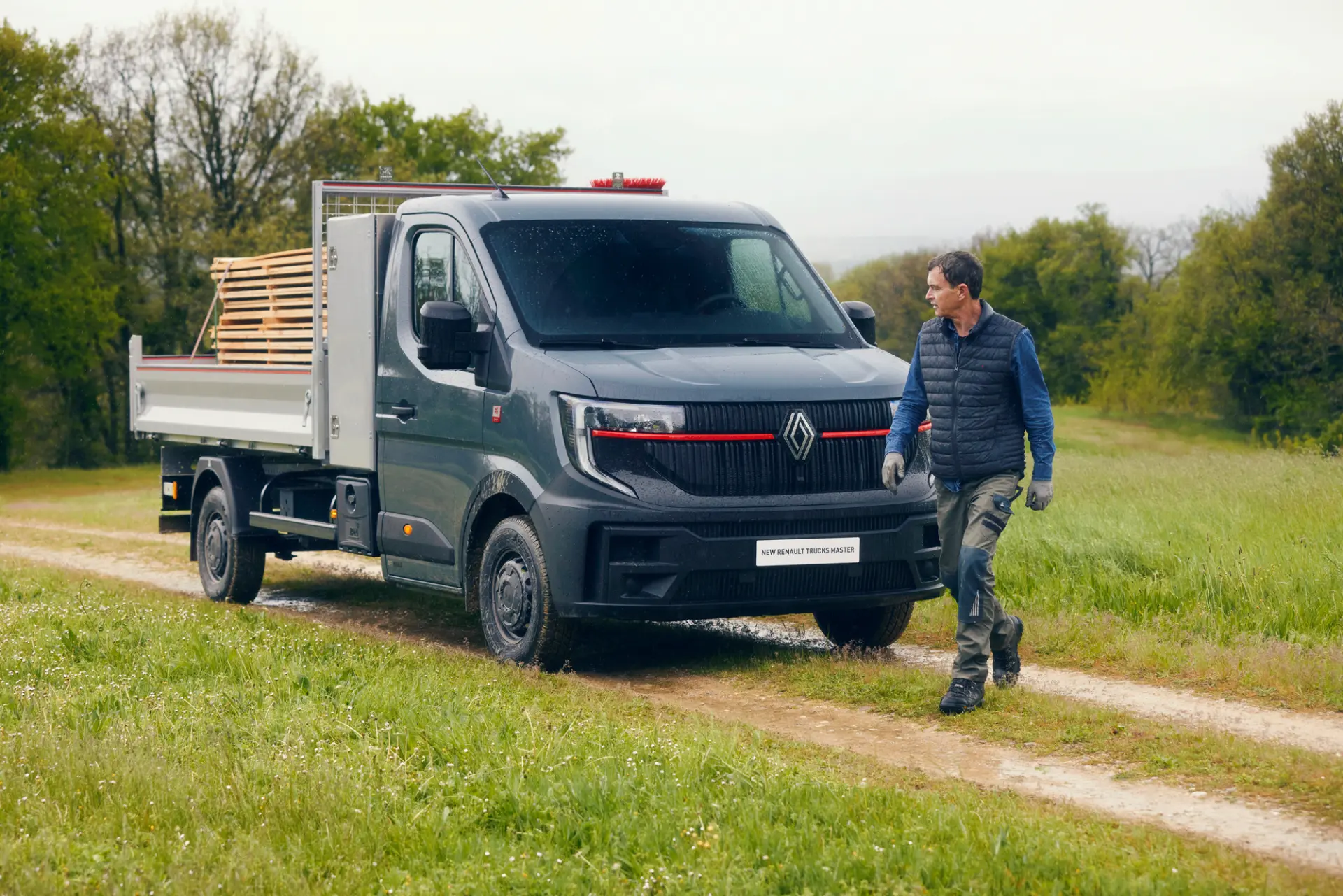 Un hombre camina junto a un camión Renault Master Red EDITION de plataforma gris que transporta tablones de madera por un camino de tierra cubierto de hierba, mostrando su capacidad como vehículo industrial de construcción.