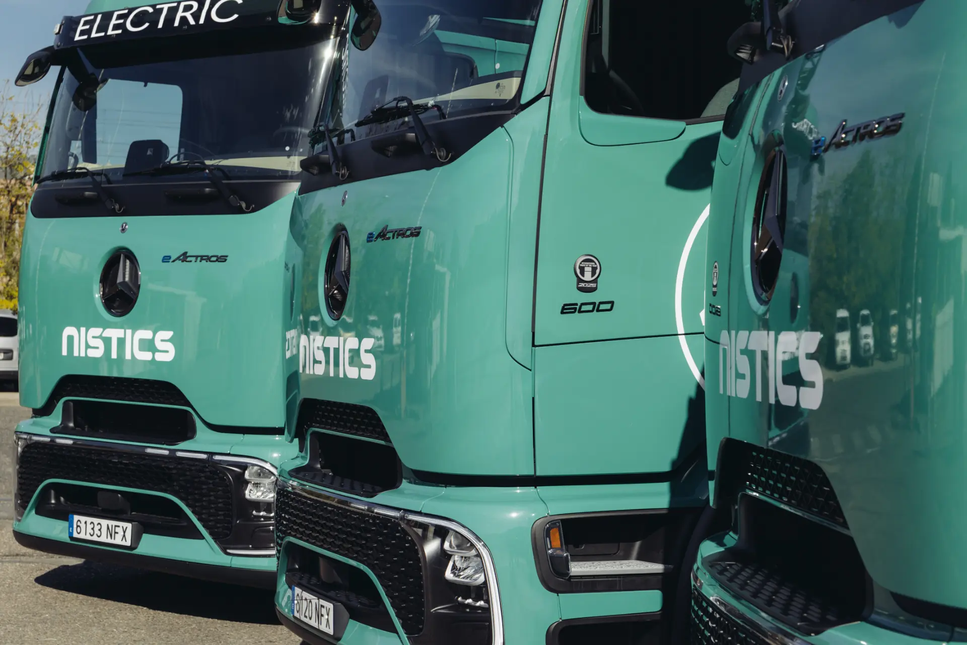 Fila de camiones de reparto eléctricos de color verde azulado con la marca "NISTICS" y el distintivo Borrador automático aparcados uno al lado del otro, con las parrillas delanteras visibles.