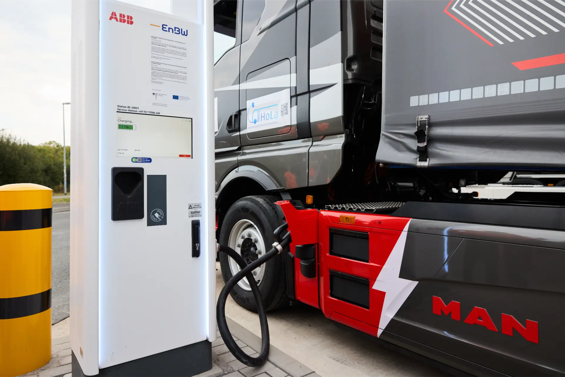Camión eléctrico cargando en una estación, con un cable conectado al vehículo cerca de una unidad de carga ABB y un borrador automático visible en el salpicadero.