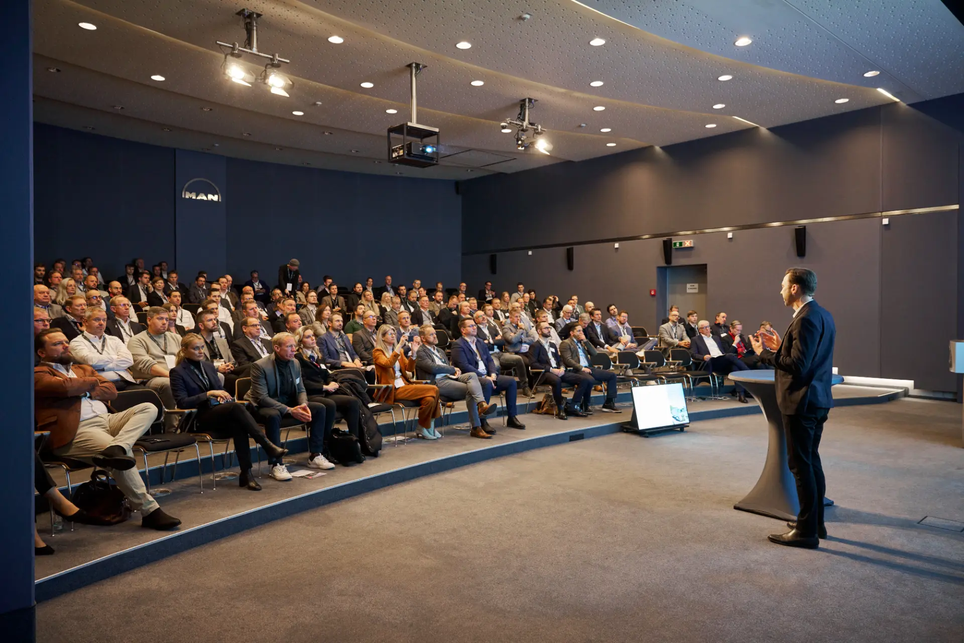 Un ponente se dirige a un público sentado en un moderno auditorio con una gran pantalla de proyección, destacando los avances en electromovilidad y las innovaciones de MAN, parte de Volkswagen Group Logistics.
