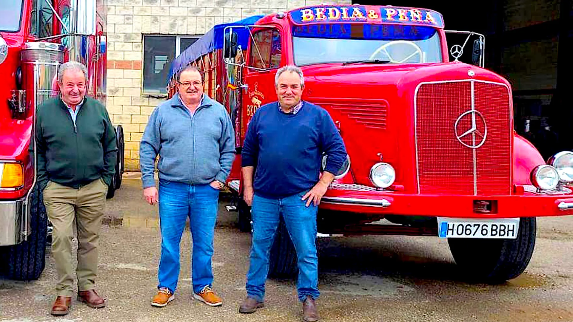 Tres hombres de pie delante de un camión rojo de época, un Mercedes Benz L 332 bellamente restaurado con la inscripción "BEDIA & PINA" en la parte delantera, muestran su pasión por los camiones clásicos restaurados.