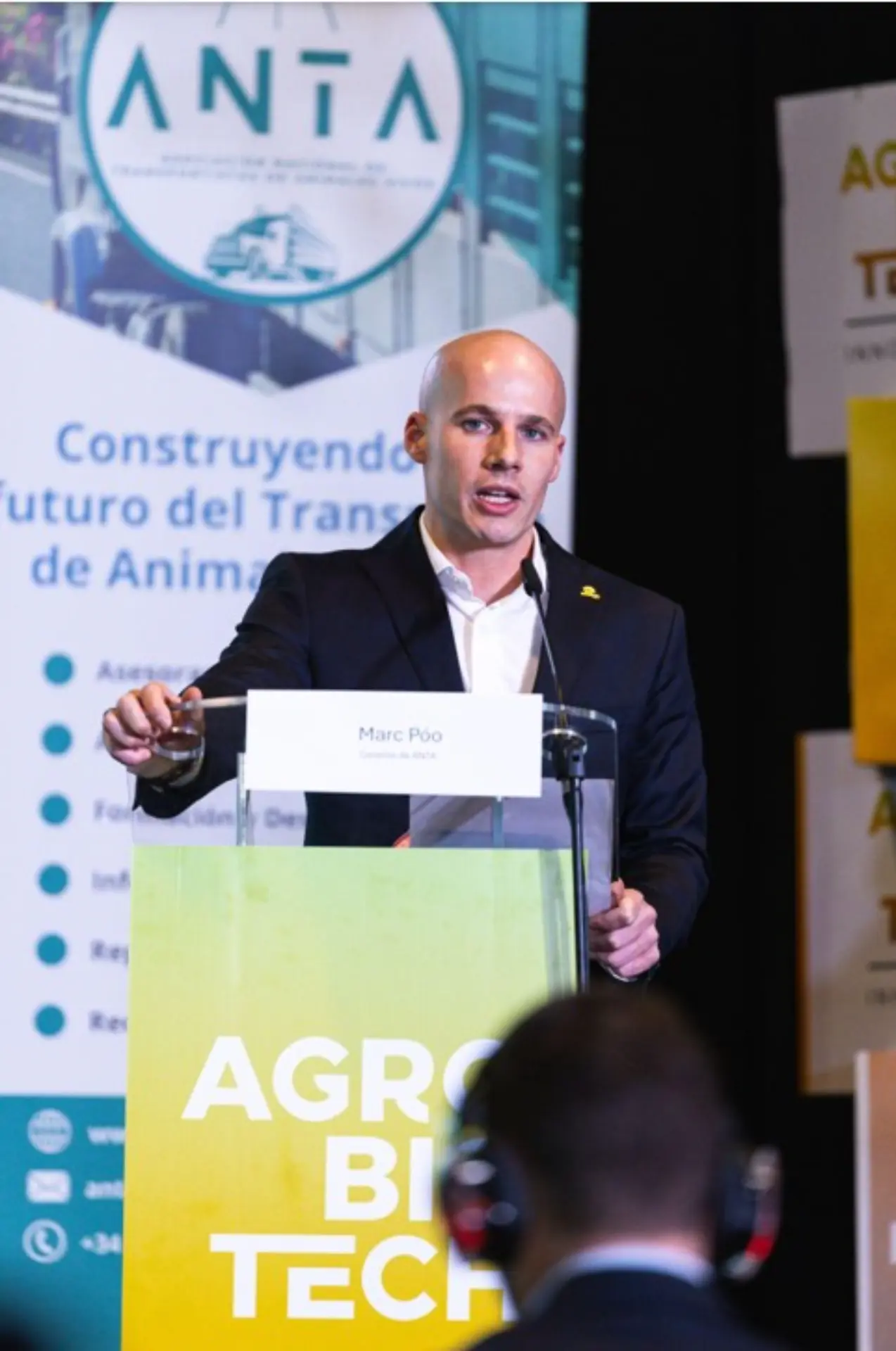 Un hombre de traje habla en un podio rotulado "AGRO BIO TECH", discutiendo el papel de ANTA en la prevención de la peste porcina en una conferencia. El nombre "Marc Pió" es visible en la parte delantera.
