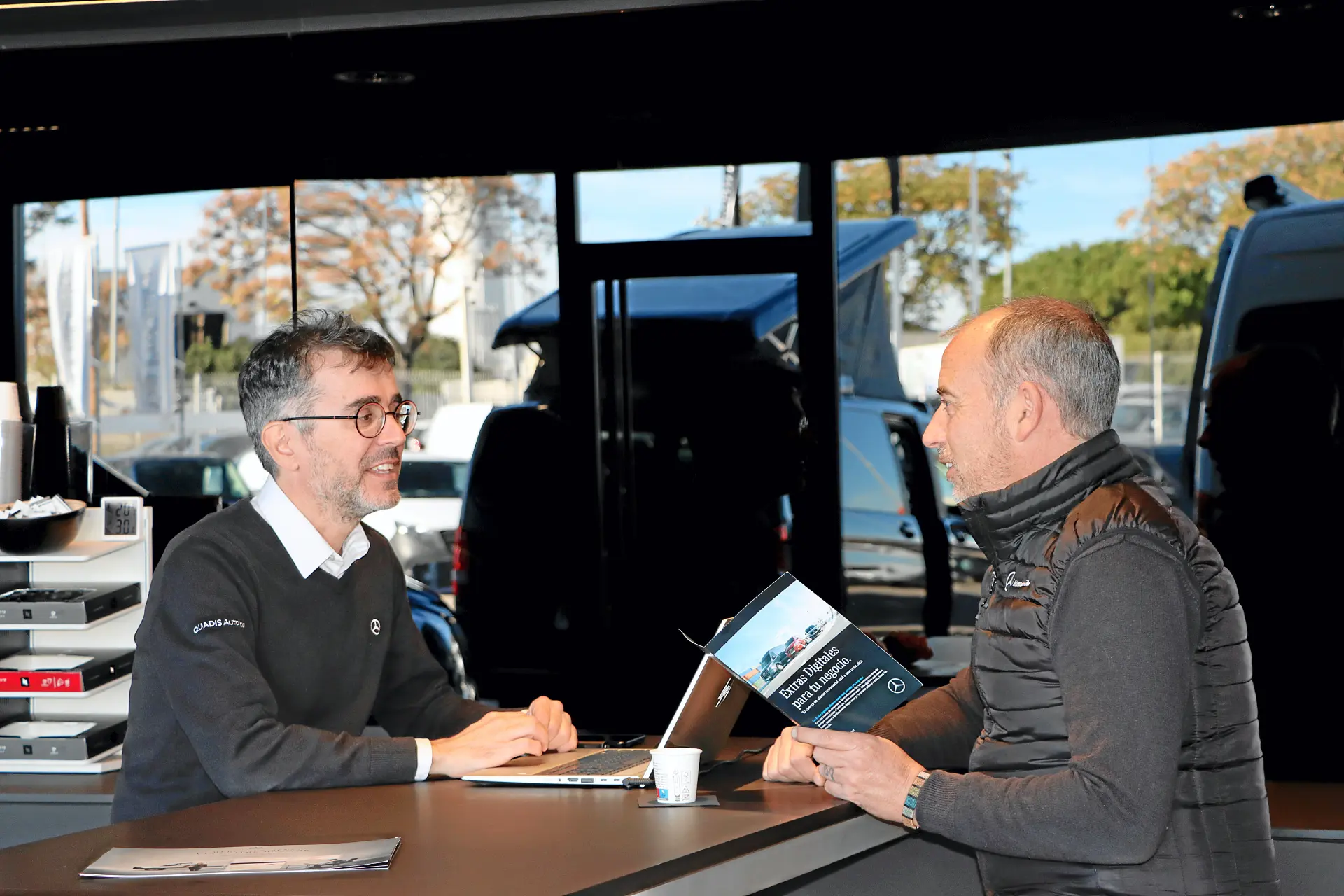 Dos hombres hablan en el mostrador de un concesionario, uno de ellos con un folleto sobre furgonetas Mercedes, con coches visibles en el exterior a través de la ventanilla, perfectos para quienes estén considerando los servicios de Talleres Autolica o Talleres Quadis.