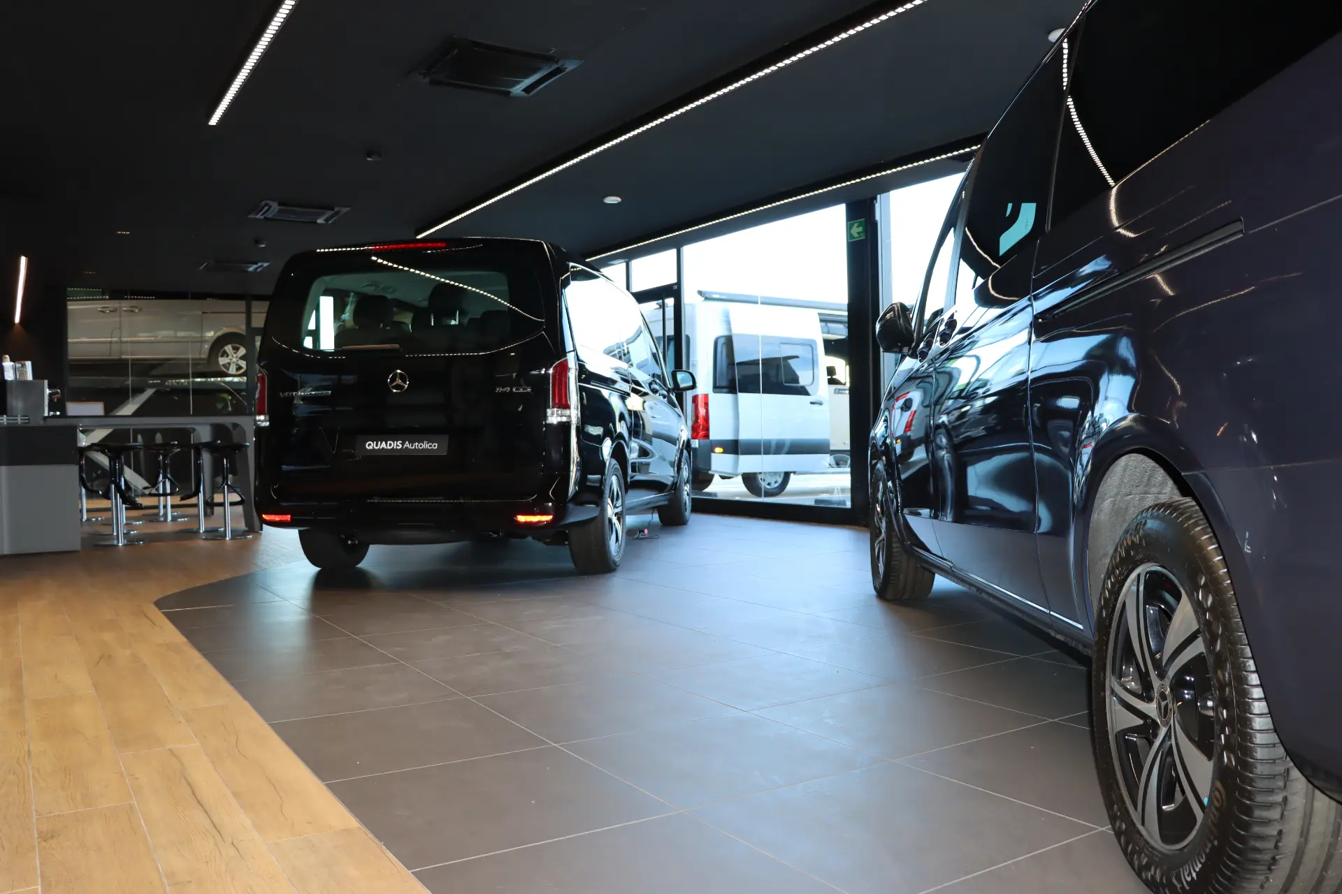 Tres Mercedes negros furgonetas se exhiben en el interior de una moderna sala de exposición de automóviles con grandes ventanales de cristal, mostrando la calidad que se espera de Quadis Autolica.