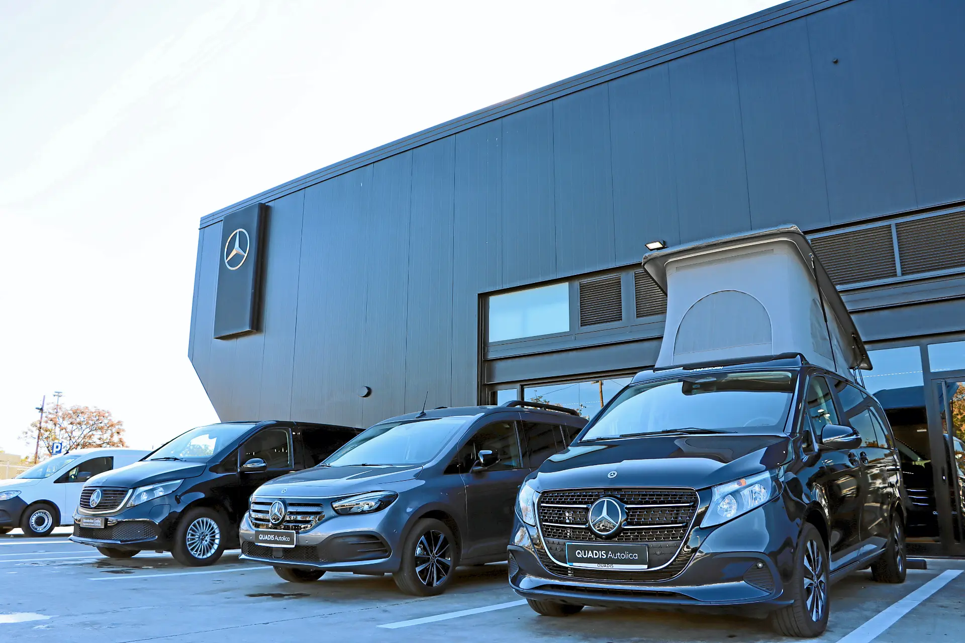 Tres furgonetas Mercedes-Benz aparcadas frente a un moderno edificio negro de concesionarios con el logotipo de Mercedes, mostrando la calidad y el servicio prestados por Quadis Autolica.