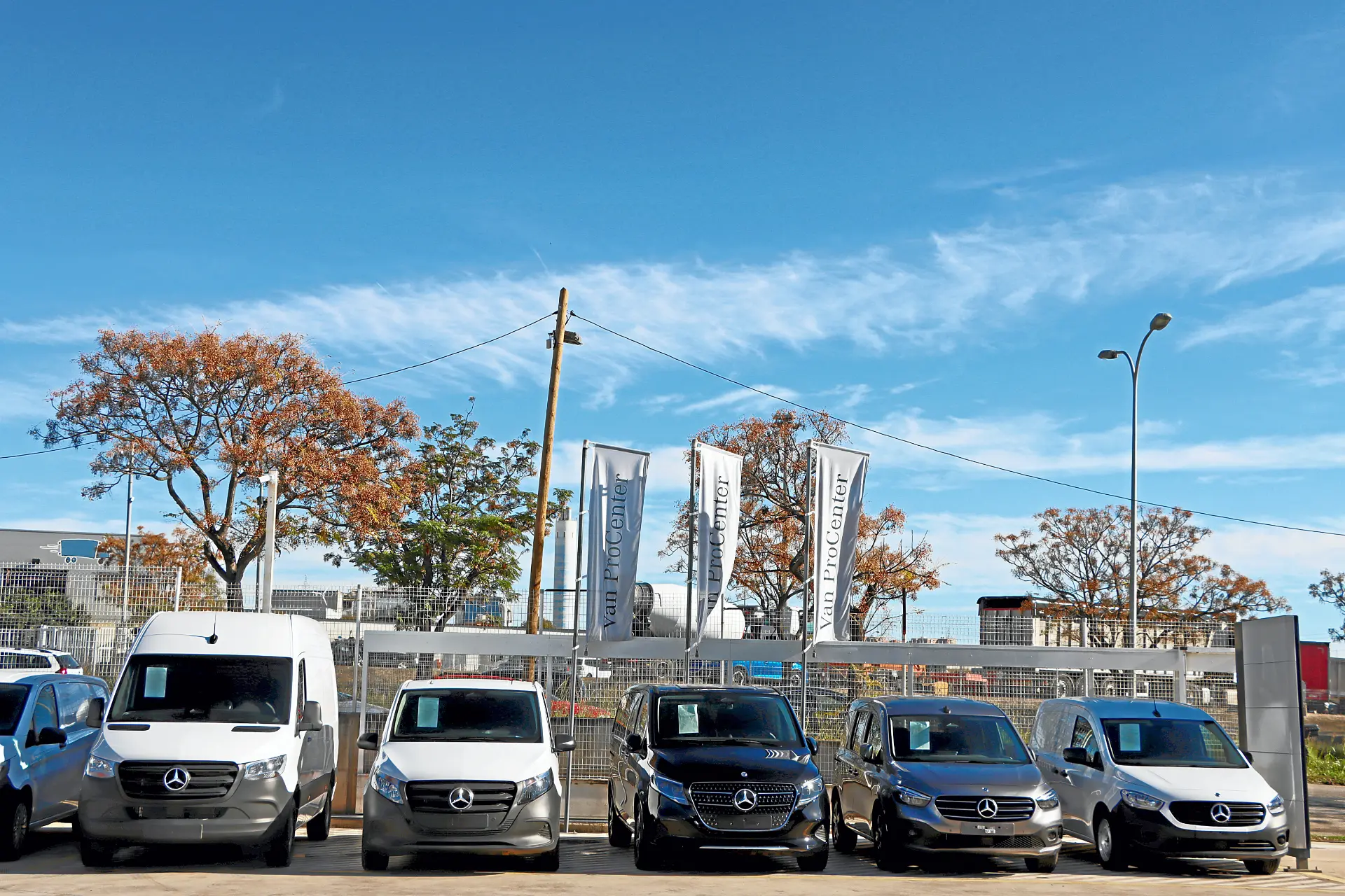 Cinco furgonetas Mercedes aparcadas en fila en un concesionario, con banderas y árboles al fondo, muestran la fiabilidad que ofrecen los talleres Autolica y Quadis Autolica.
