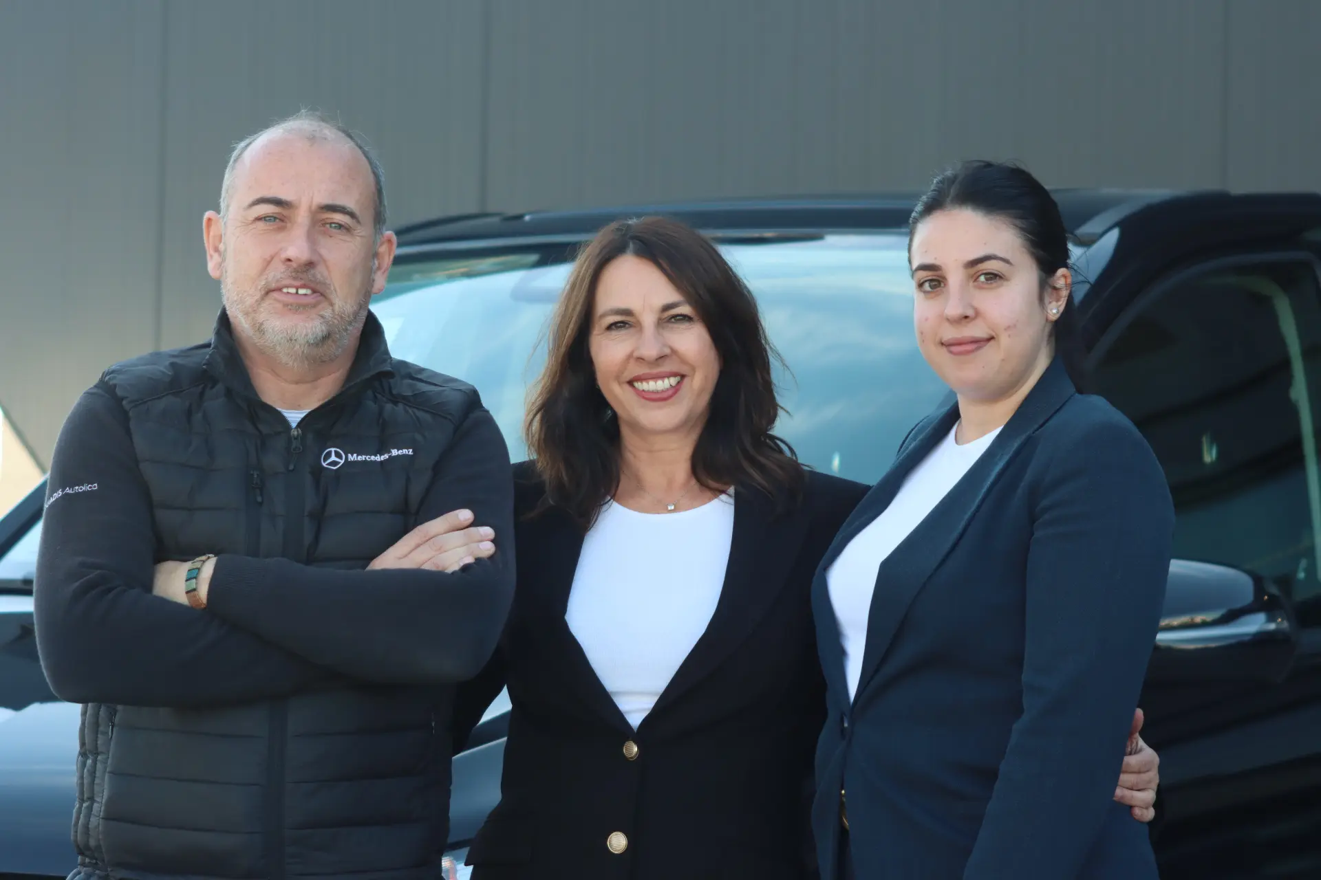 Tres personas en traje de negocios sonríen mientras están de pie delante de un coche negro al aire libre, destacando el servicio profesional en Talleres Quadis.
