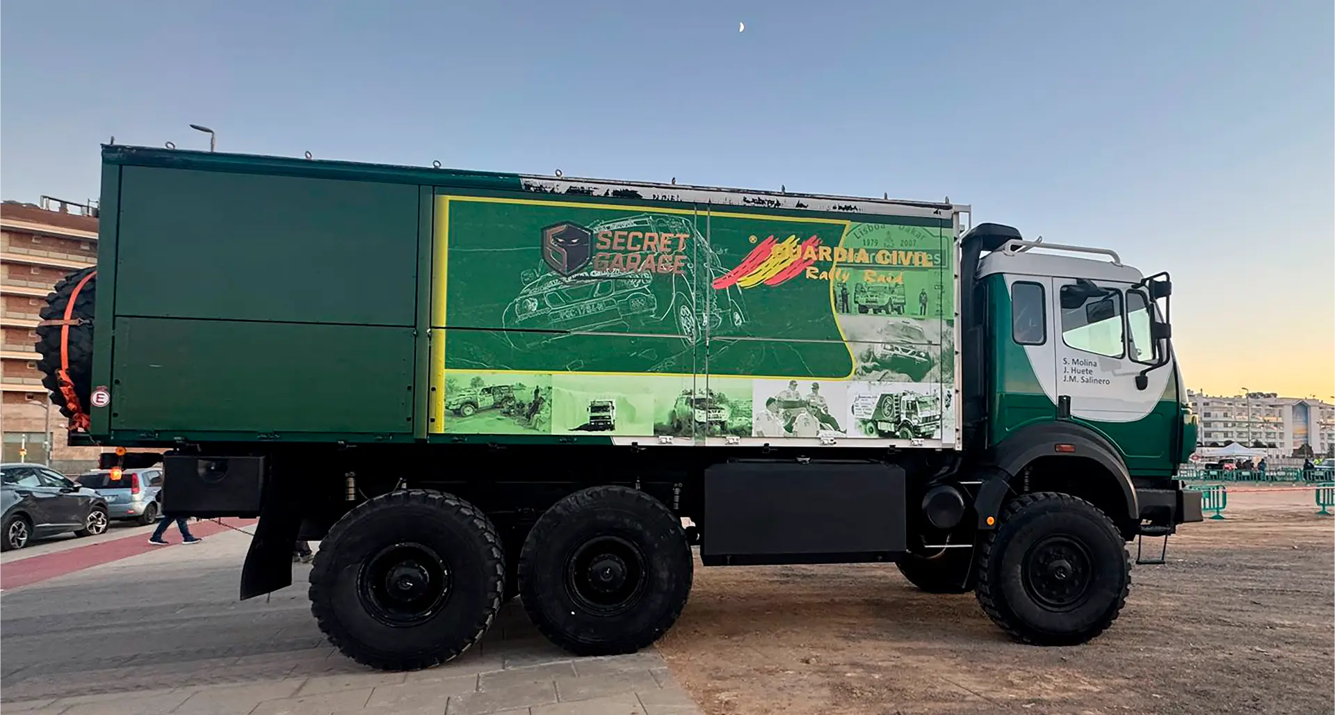 Gran camión verde 6x6 con un cartel de "Atlas Secreto" y "Guardia Civil", que recuerda a los rallies del Dakar, aparcado en una calle de la ciudad al anochecer.
