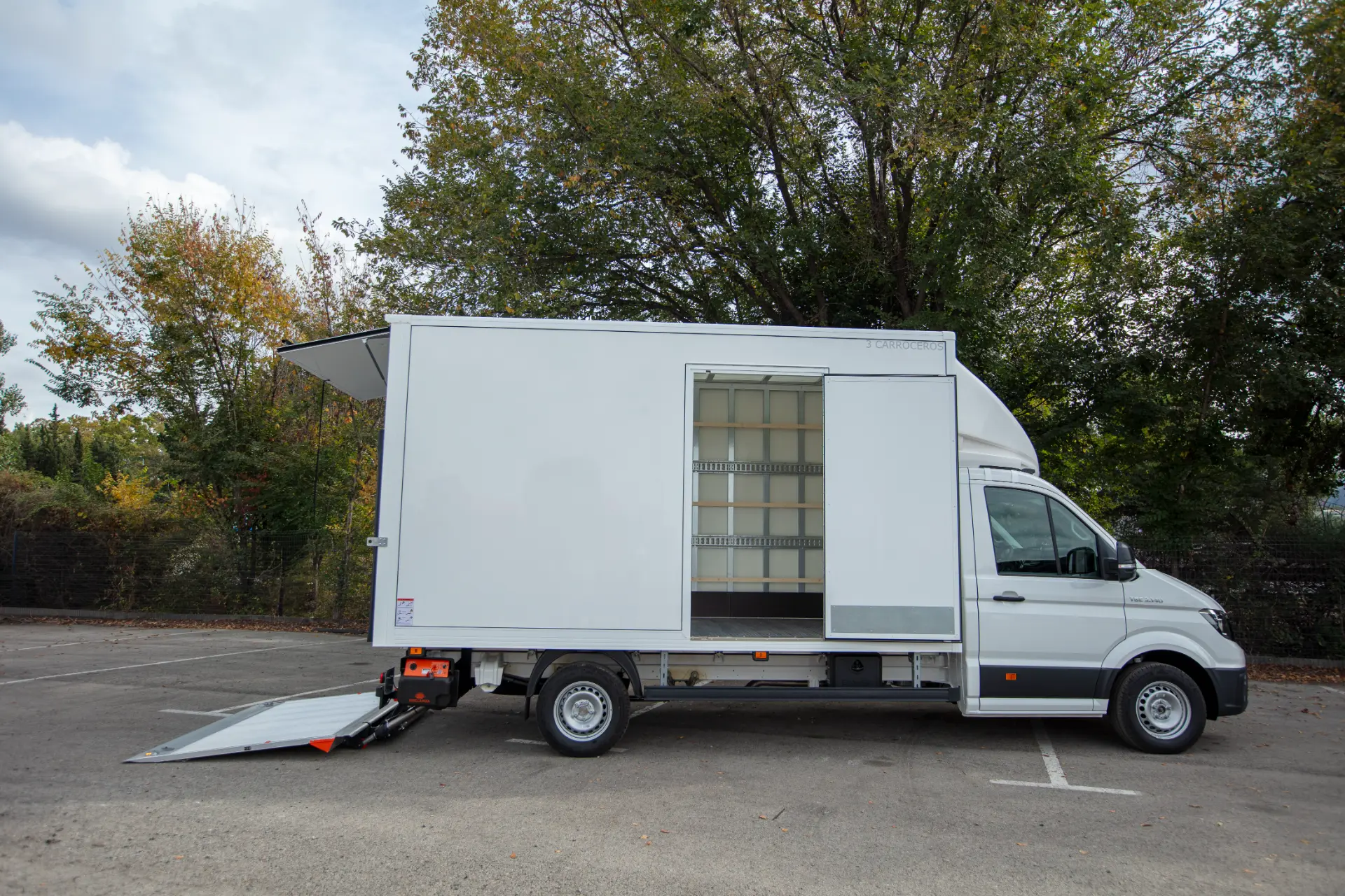 Un camión de reparto blanco con una puerta lateral abierta y una rampa de carga trasera se encuentra en un aparcamiento cerca de árboles, listo para realizar eficientes operaciones de borrador automático.