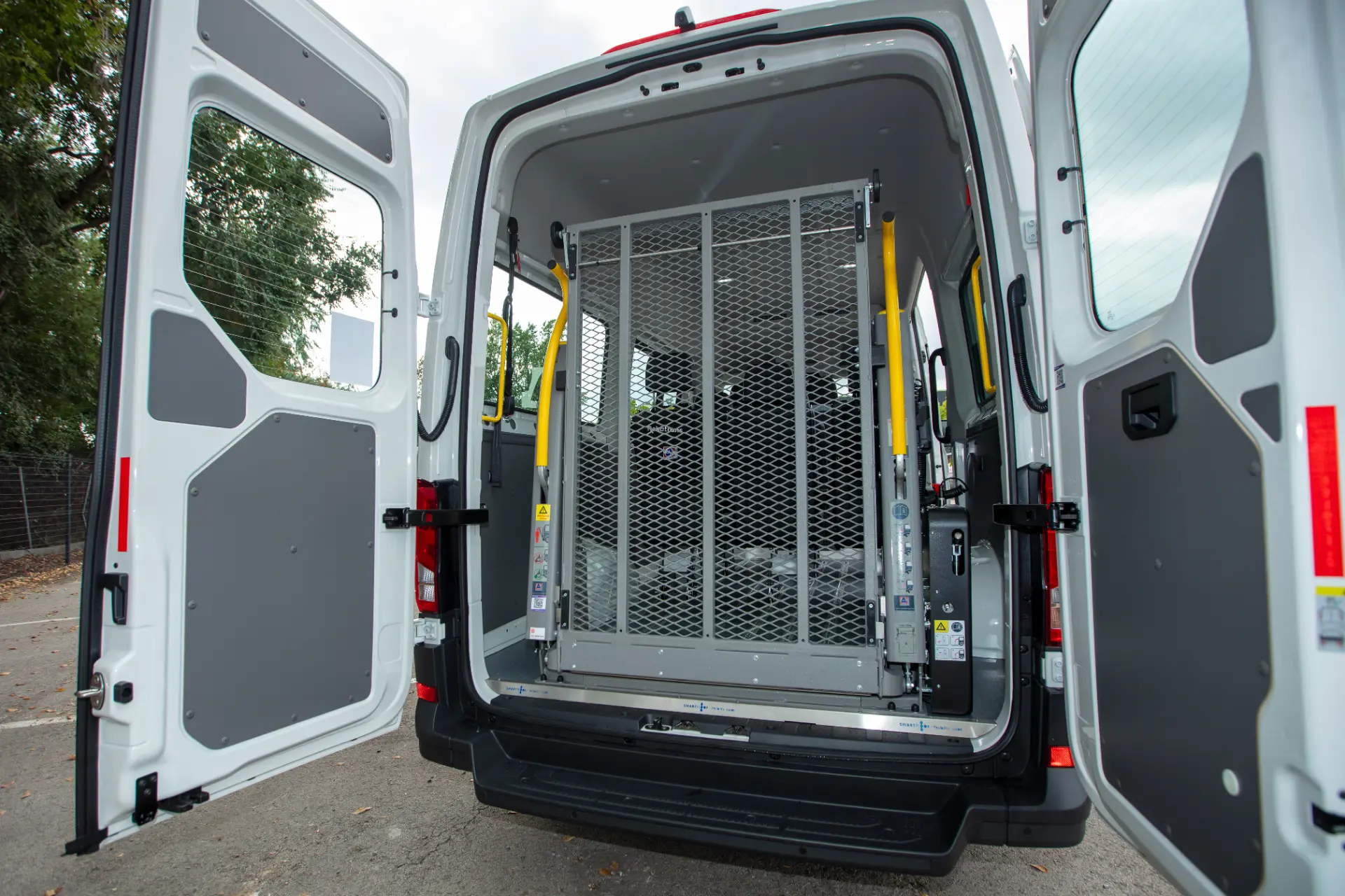Puertas traseras abiertas de una furgoneta que muestran un elevador metálico para sillas de ruedas, barandillas de seguridad amarillas y un sistema Borrador automático en el interior.