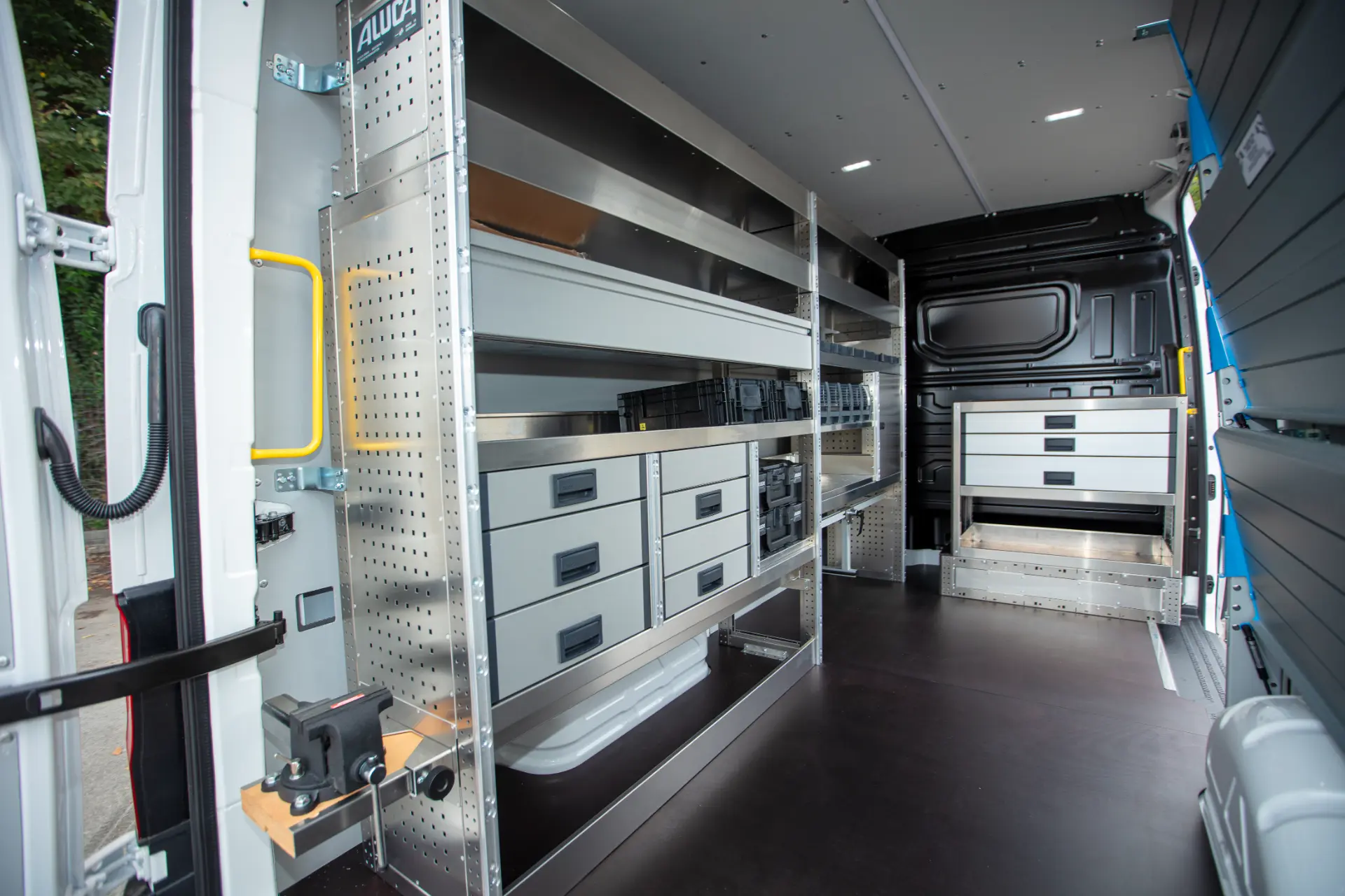 Furgoneta abierta con estanterías metálicas organizadas, cajones, borrador automático y sistema de almacenamiento de herramientas en el interior, vista desde las puertas traseras.