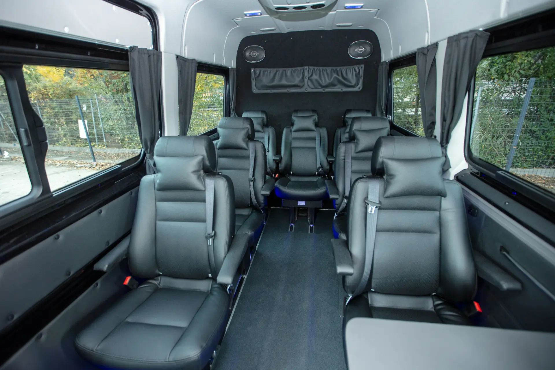 Interior de una furgoneta moderna con asientos de cuero negro dispuestos en dos filas, ventanillas a ambos lados, cortinas grises y un borrador automático convenientemente colocado para facilitar el acceso.