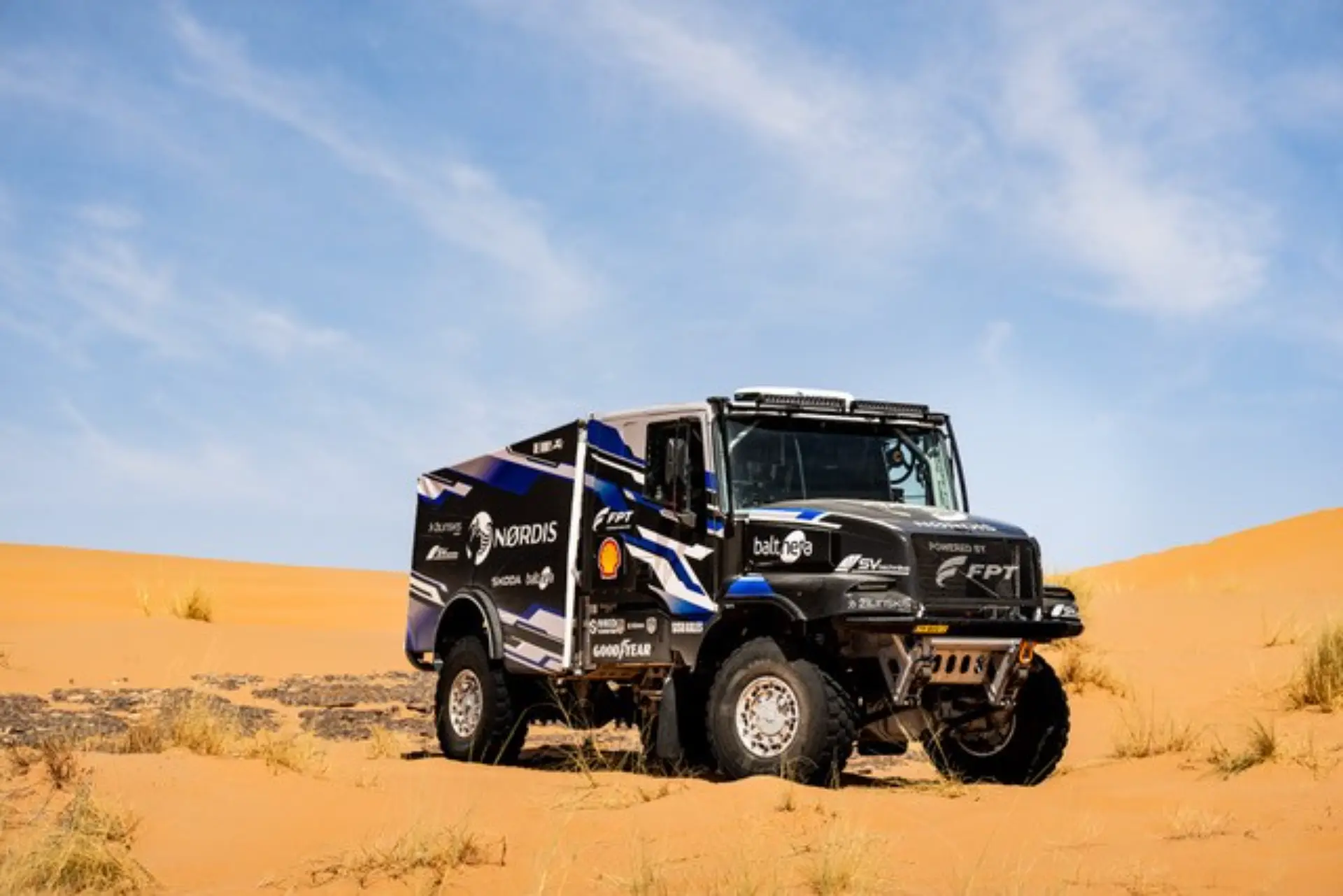 Un camión de rally con logotipos de patrocinadores y una calcomanía de "Borrador automático" circula por la arena naranja del desierto bajo un cielo azul.