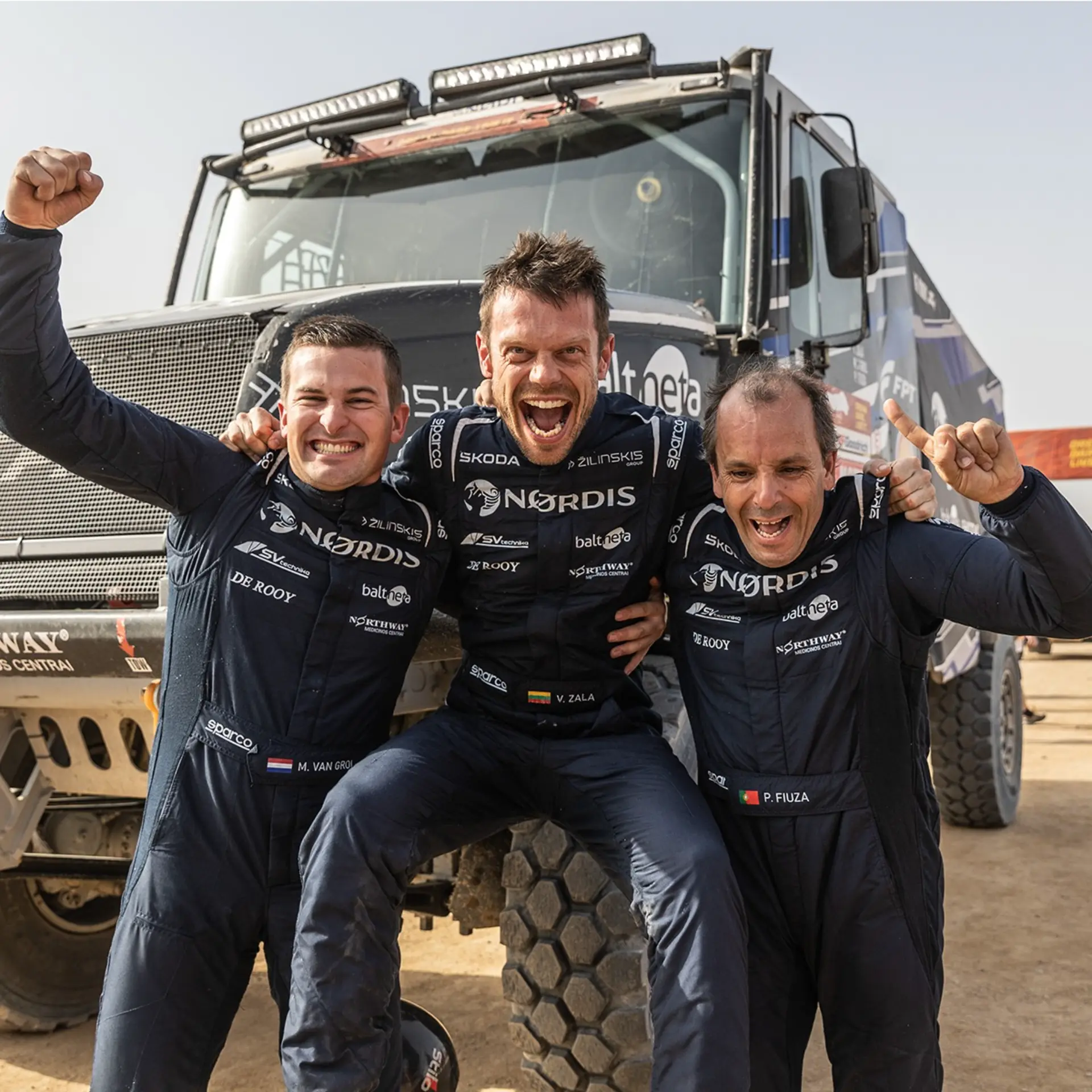 Tres jubilosos pilotos de carreras del Team De Rooy celebran delante de un gran camión de rally, sonriendo y levantando el puño.