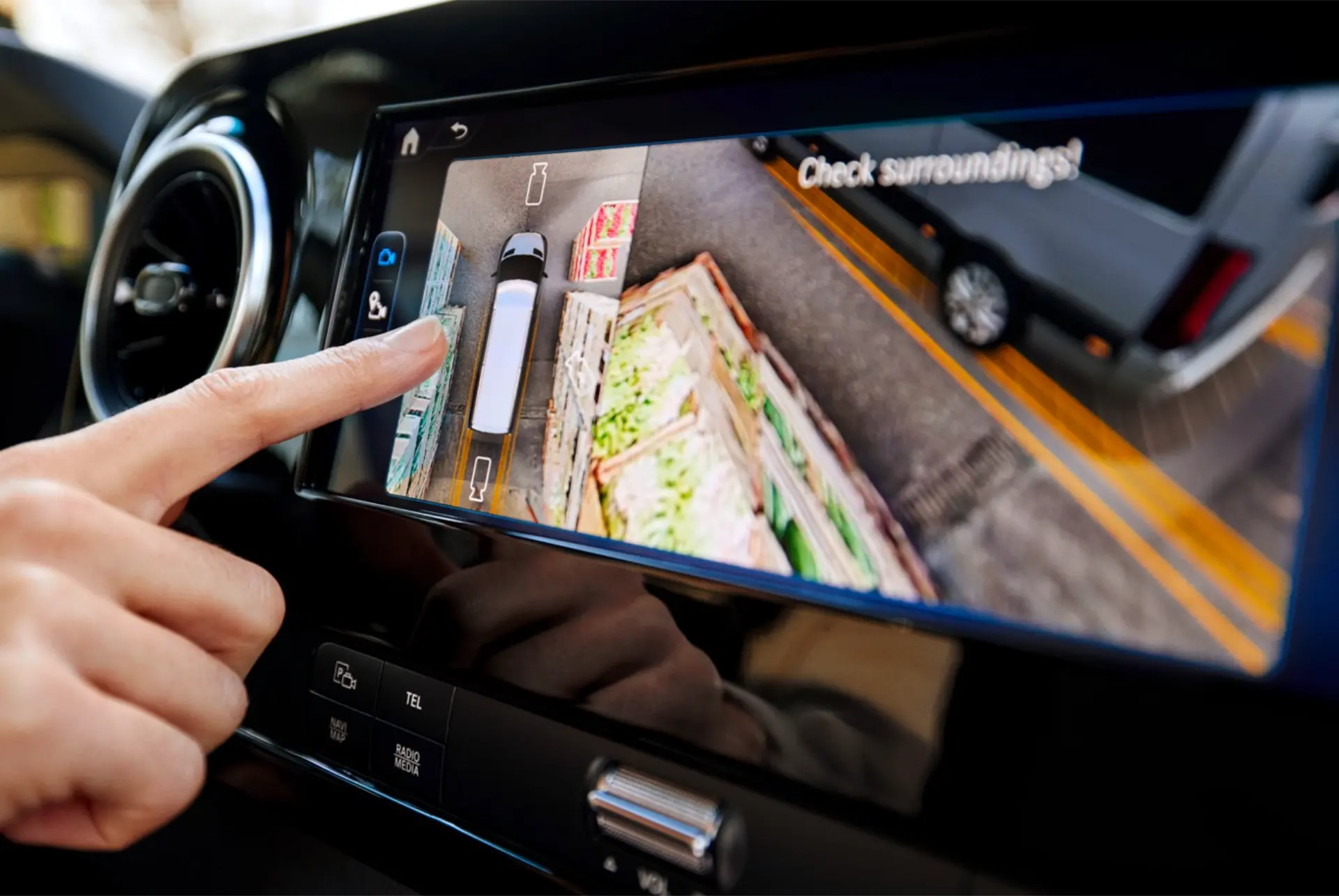 Una mano toca la pantalla táctil del Mercedes-Benz eSprinter que muestra una cámara de visión trasera con líneas de guiado para aparcar cerca de las cajas de la compra, ideal para profesionales que necesitan precisión en cada maniobra.