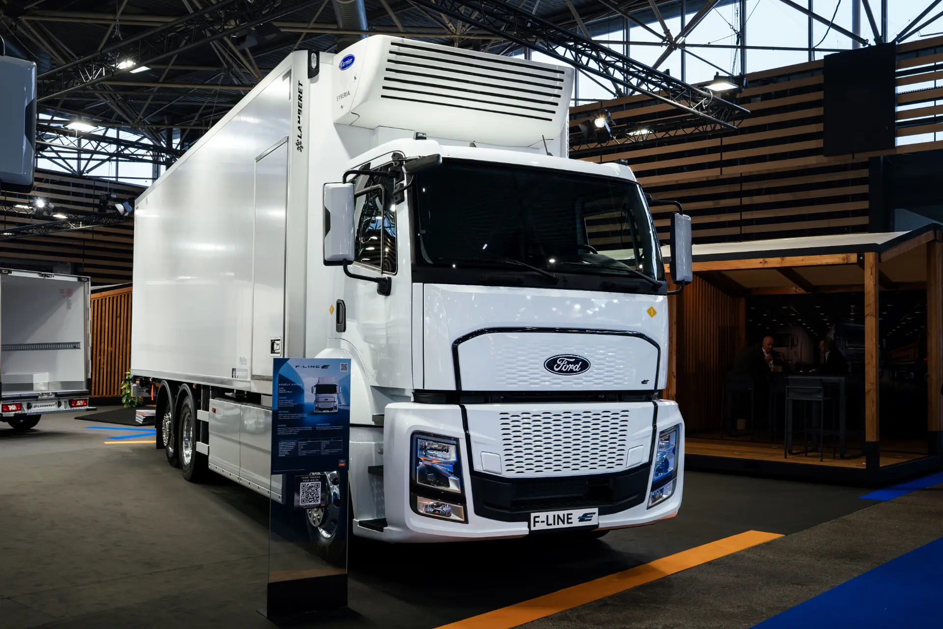 Un gran camión blanco Ford Trucks F-LINE E eléctrico se exhibe en el interior bajo brillantes luces de exposición, representando el compromiso con la logística de cero emisiones.