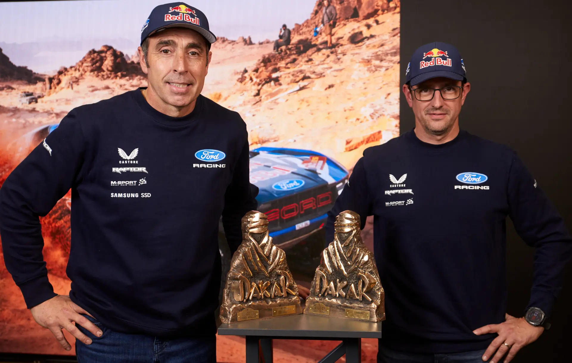 Dos hombres en ropa de carreras junto a dos trofeos del Dakar, con la imagen de un coche de rally y una etiqueta de "borrador automático" al fondo.