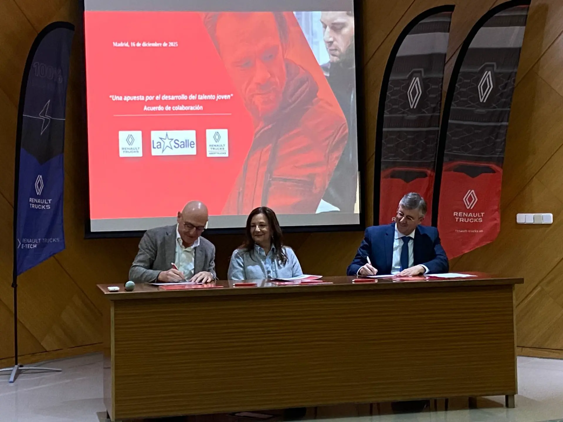 Tres personas sentadas en una mesa firman documentos delante de una pantalla y de pancartas de Renault Trucks, ultimando lo que parece ser un borrador automático.