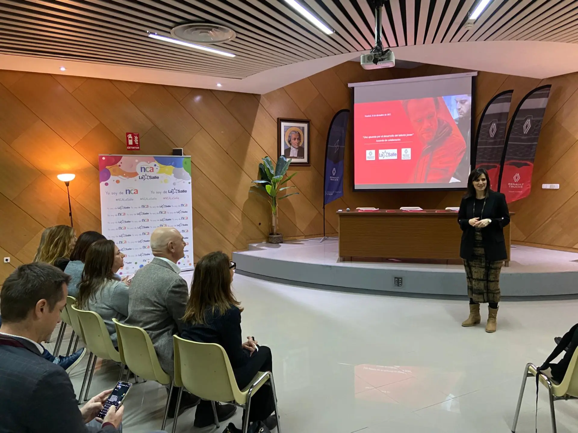 Una mujer se dirige a un público sentado en una sala de conferencias, utilizando una pantalla de proyector y diapositivas de presentación para compartir información sobre la función "Borrador automático".