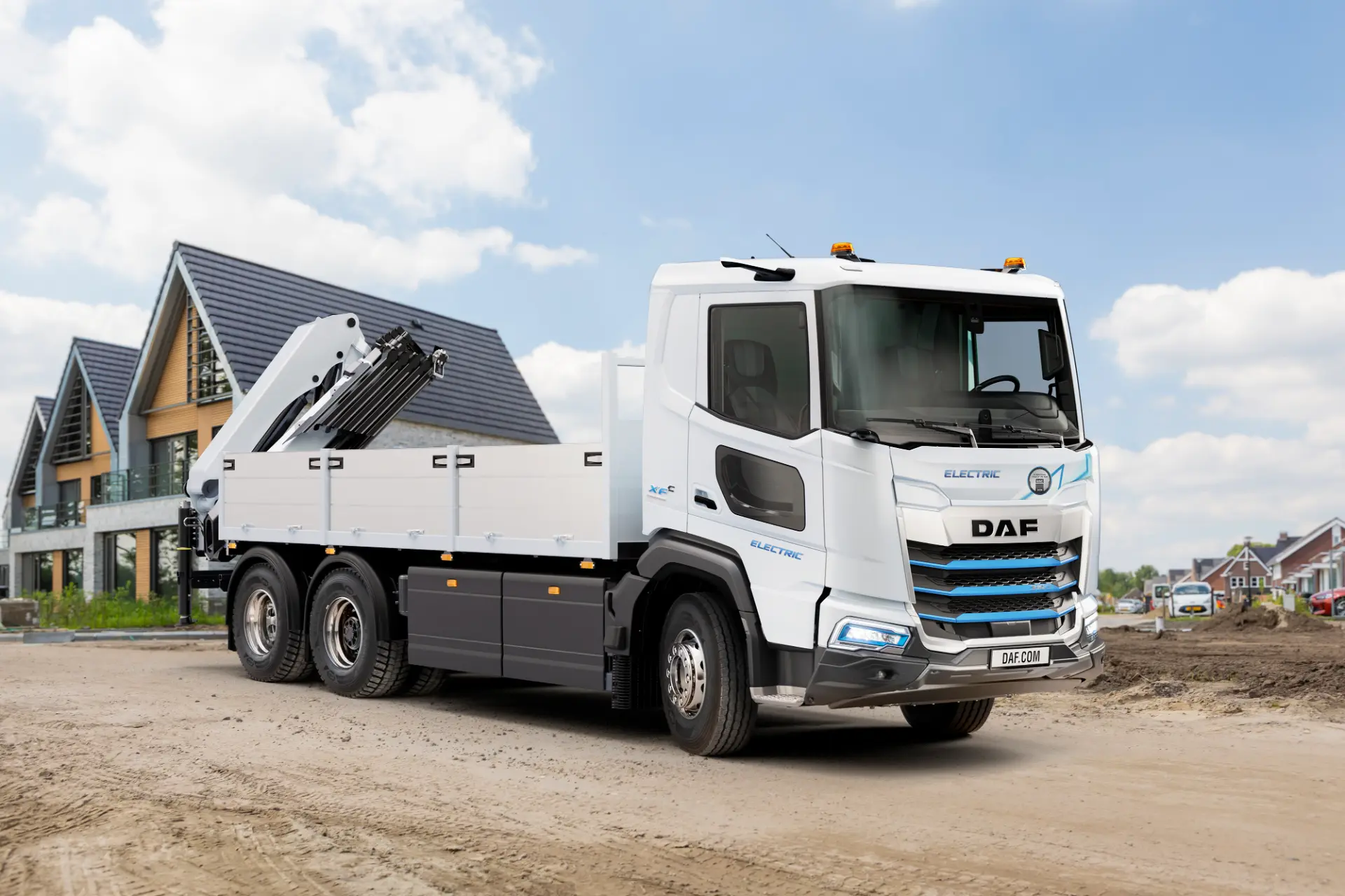 Camión DAF blanco de plataforma eléctrica con grúa, dotado de un sistema Borrador automático, estacionado en un camino de tierra cerca de casas en construcción.