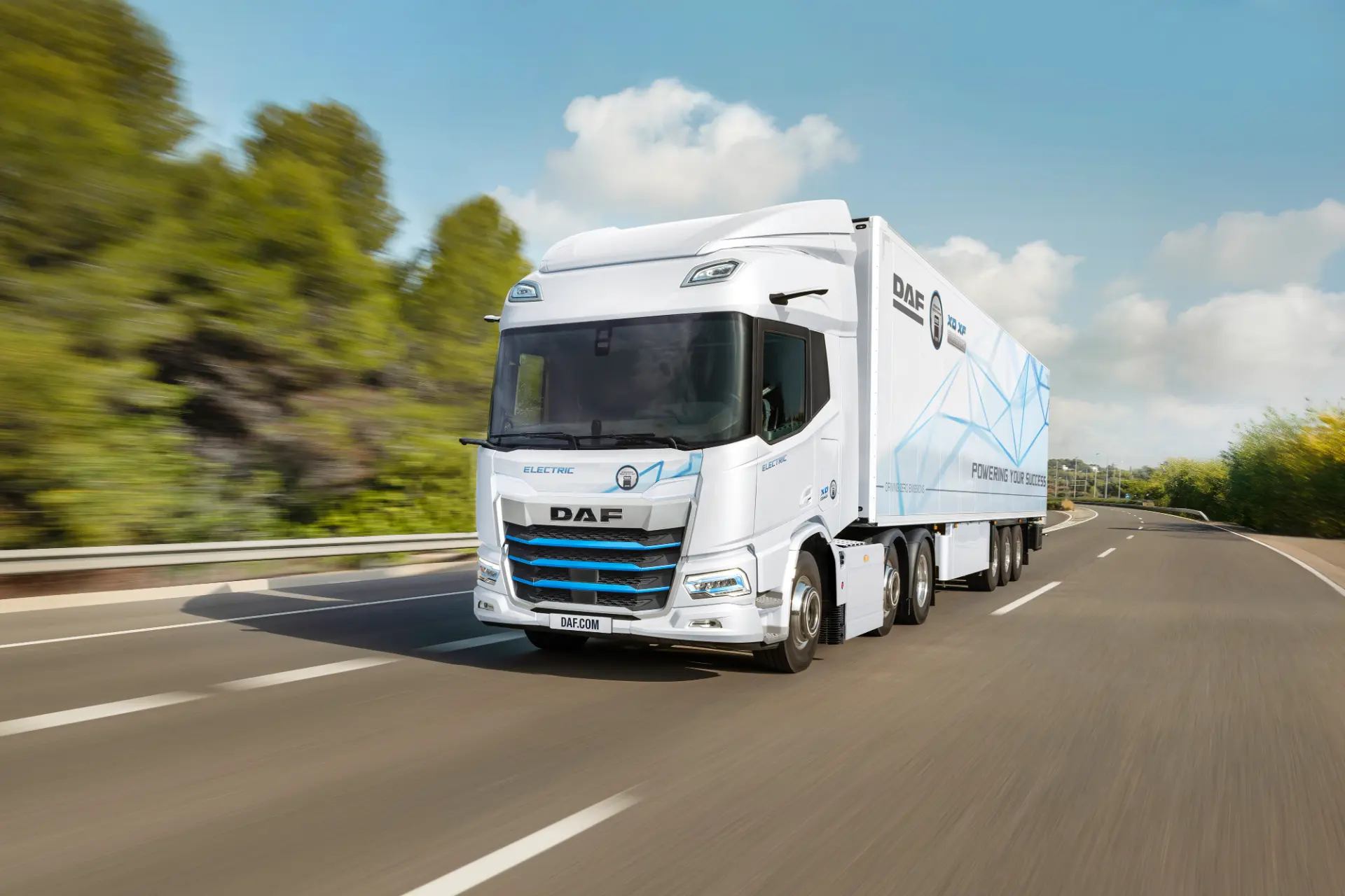 Un camión eléctrico DAF blanco con borrador automático circula por una autopista rodeada de árboles y un cielo azul despejado.