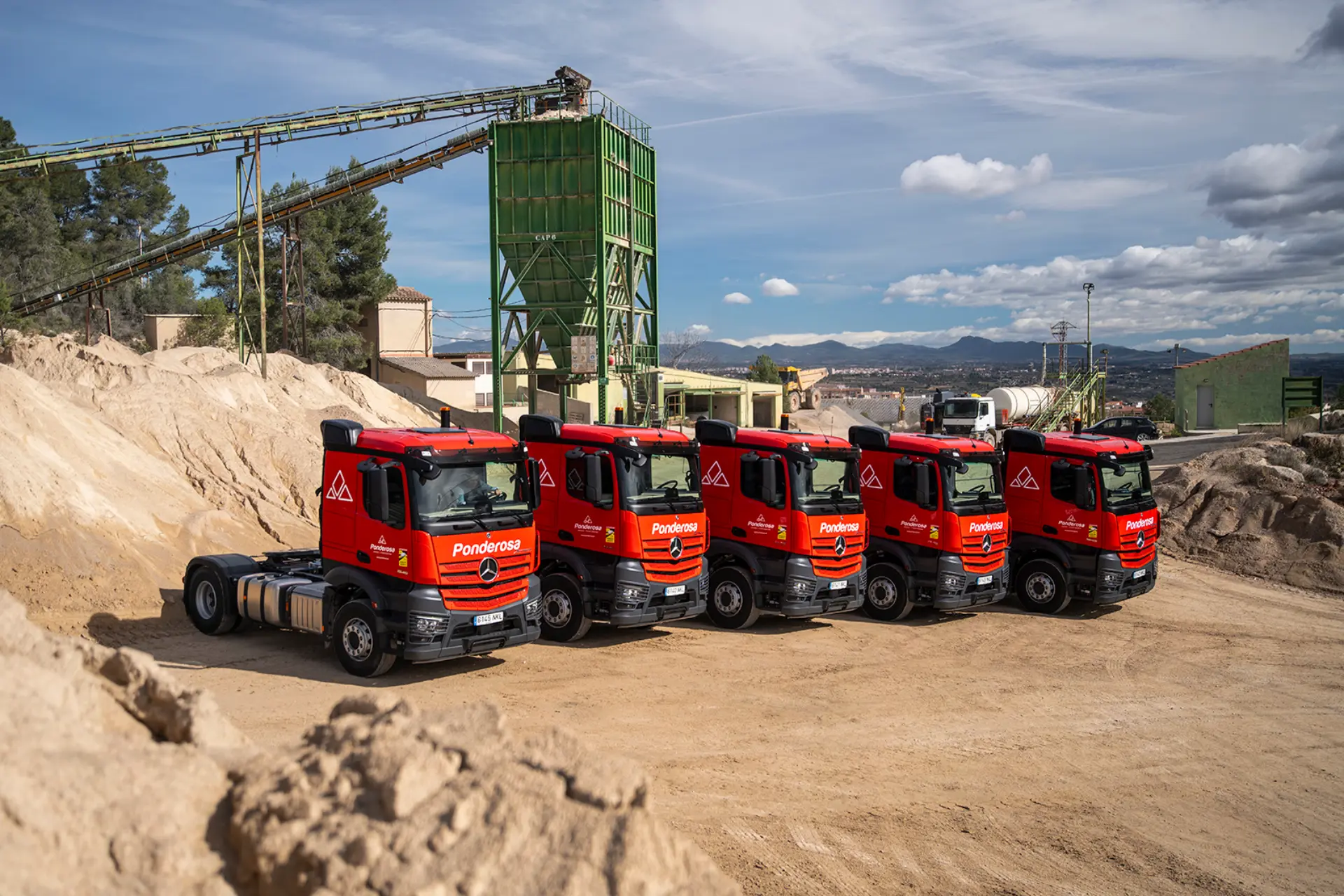 Cinco camiones rojos Pandetrans Mercedes-Benz Actros aparcados en fila en un polígono industrial de Canteras La Ponderosa, con maquinaria al fondo.
