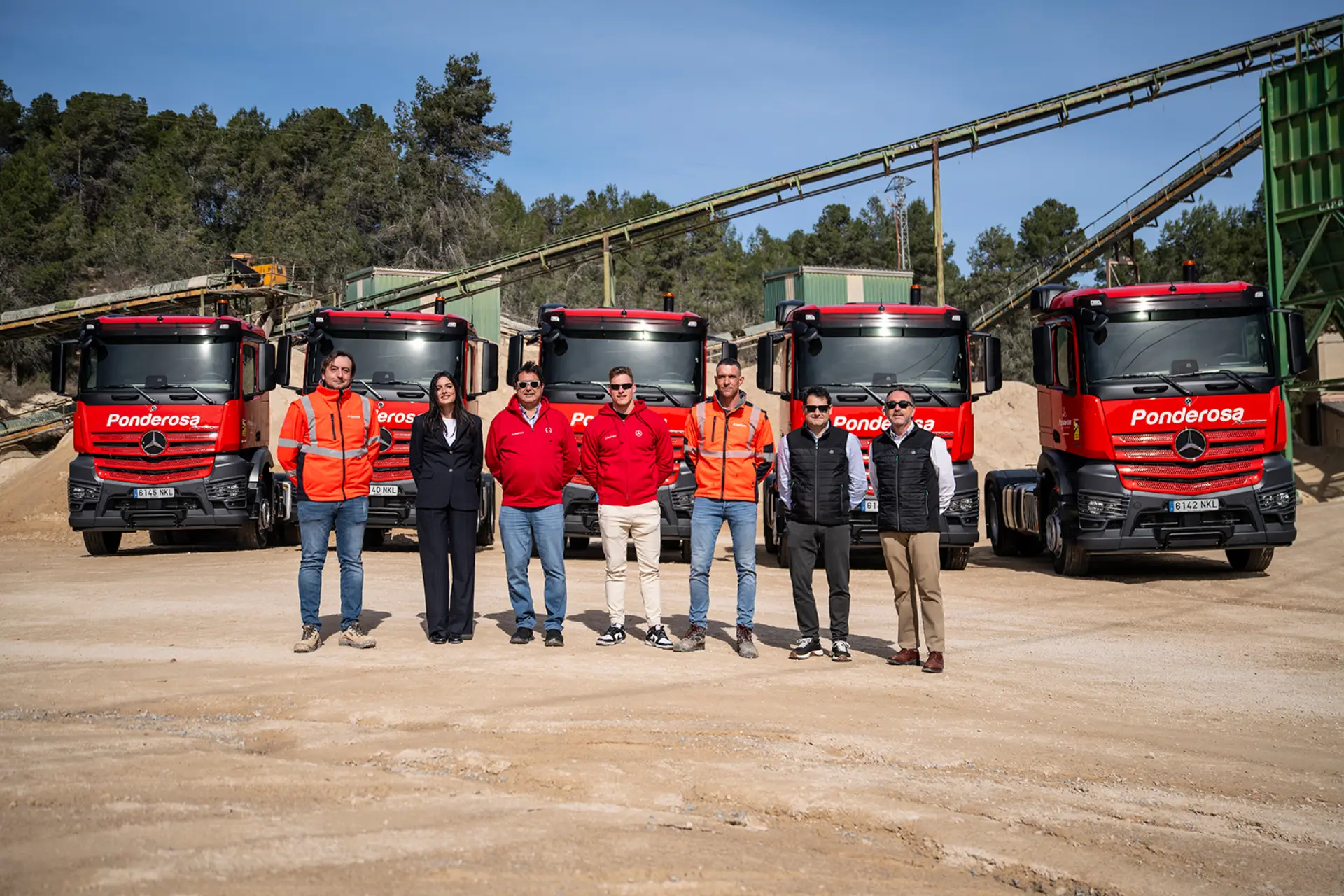 Siete personas de pie delante de cinco camiones rojos de Canteras La Ponderosa, entre ellos modelos Mercedes-Benz Actros, en una obra con árboles y equipos.
