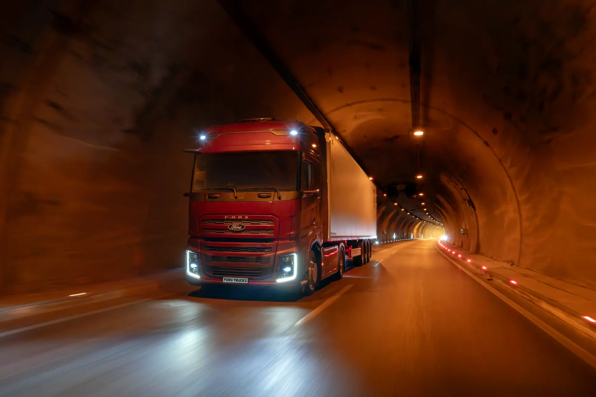 Un semirremolque rojo atraviesa un túnel bien iluminado con paredes y luces superiores anaranjadas.