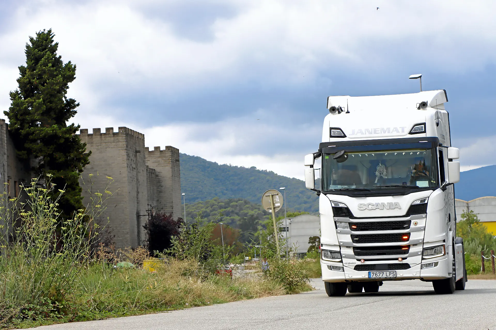 Un camión Scania blanco circula por una carretera cerca de un muro de piedra y árboles verdes bajo un cielo nublado, mostrando el viaje de Janet Torres de Soy Camionera.